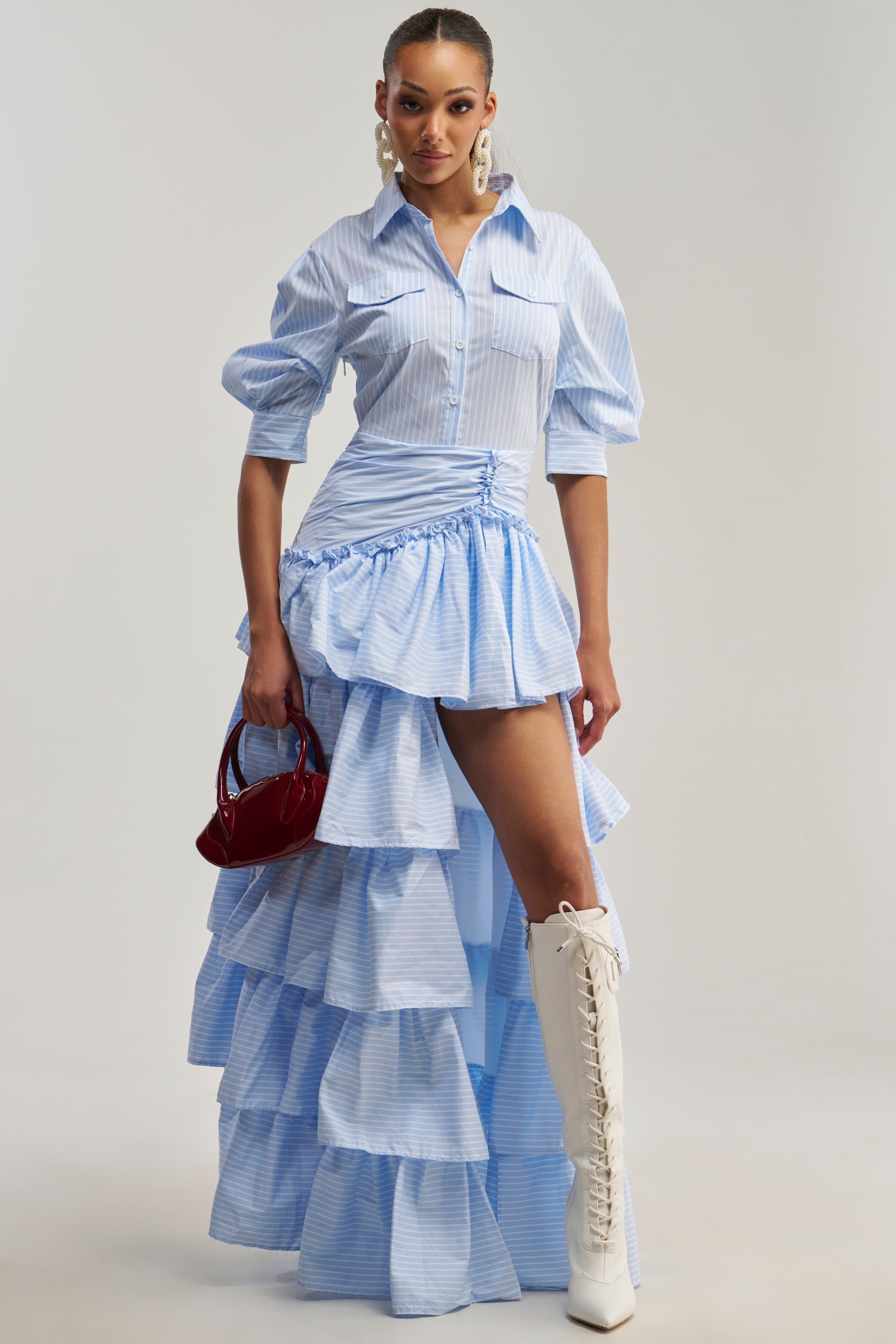 Wearing the IT’S A DATE POPLIN MAXI DRESS IN BLUE with its ruffled high-low hem, white lace-up knee-high boots, statement earrings, and a small red handbag, she poses elegantly against a plain light background—effortlessly styled for brunch.