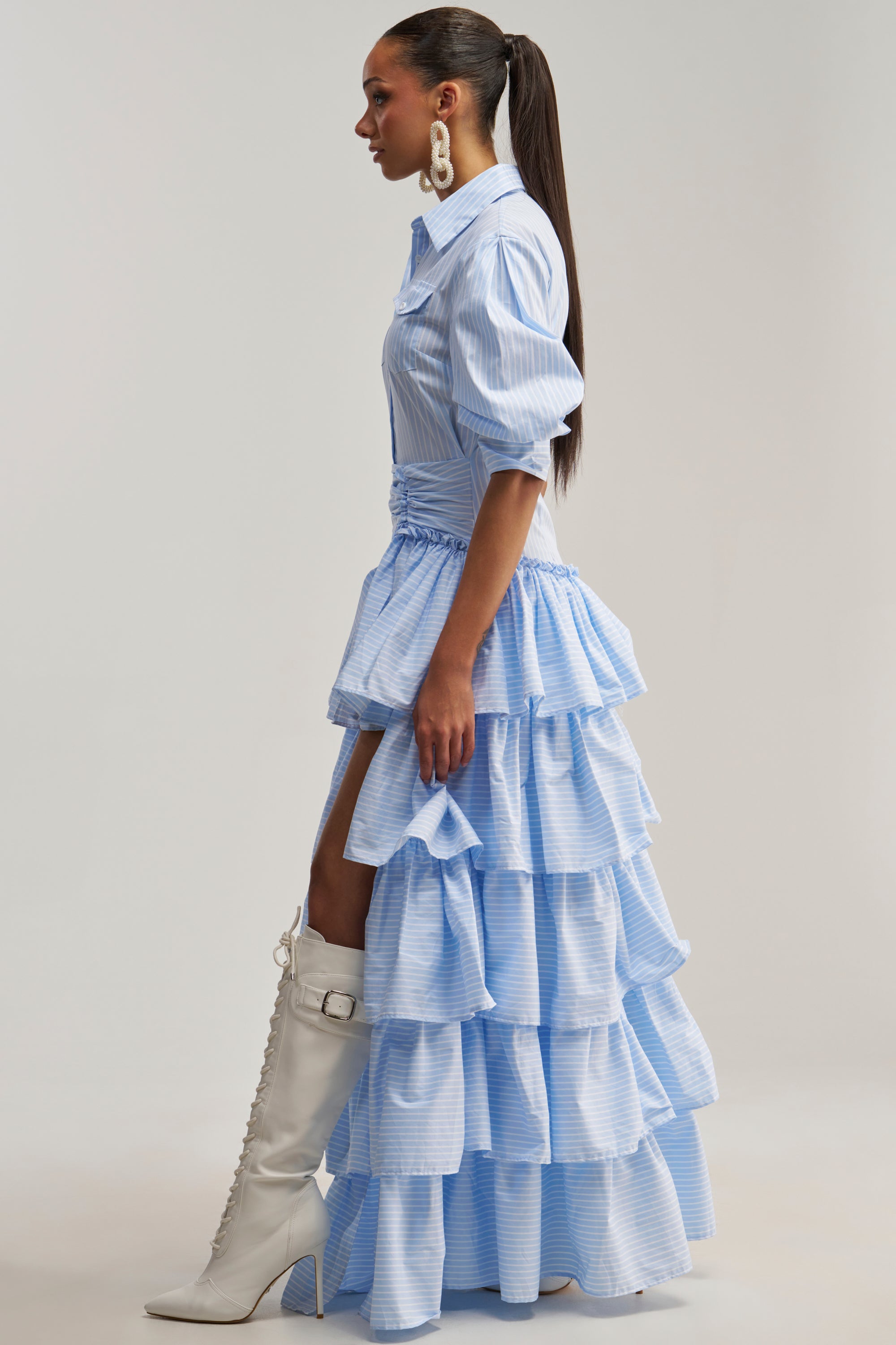 Woman in profile wearing the IT’S A DATE POPLIN MAXI DRESS IN BLUE, styled with puffed sleeves and ruffles, paired with white tall boots and large statement earrings, standing against a plain background.