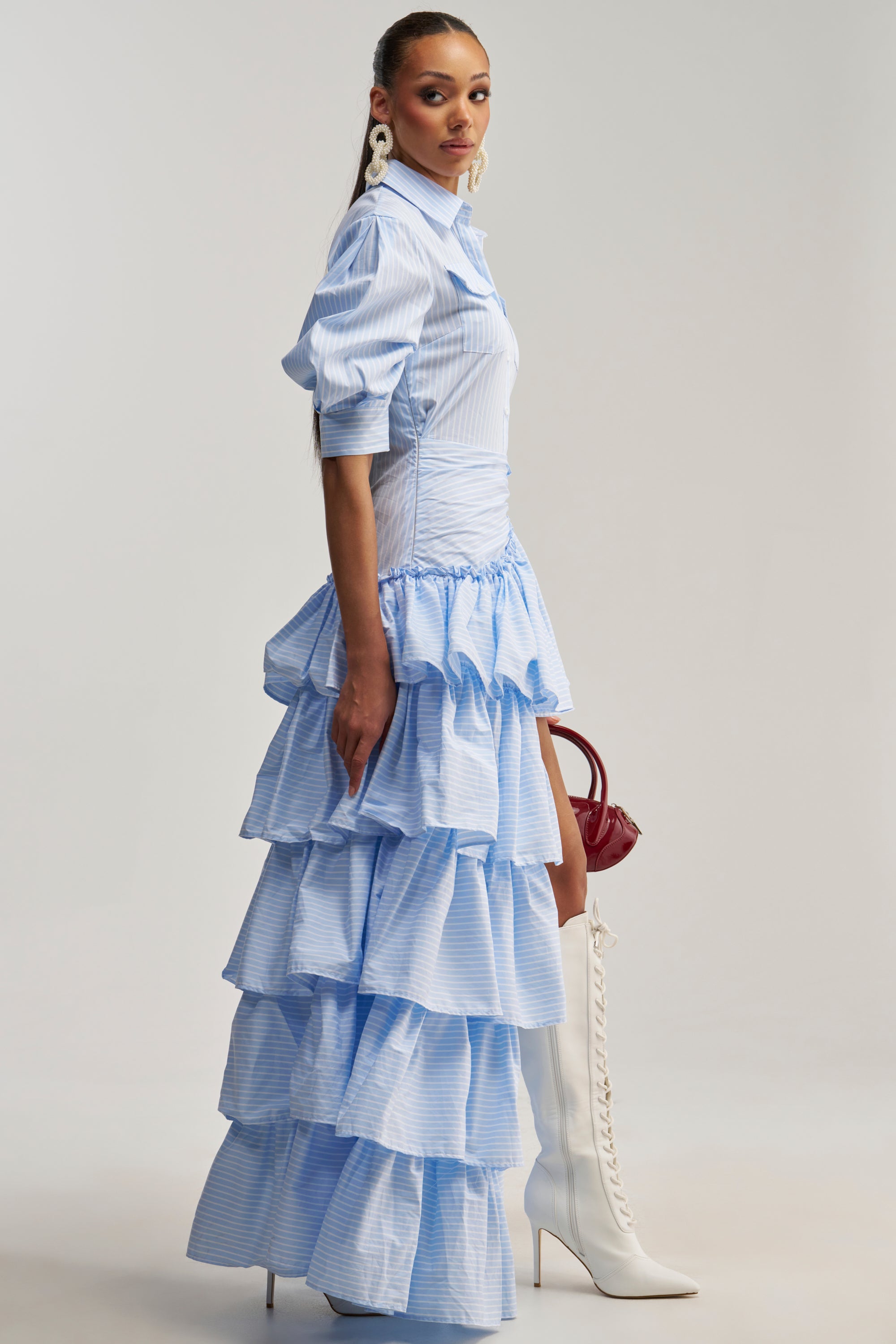 Wearing the IT’S A DATE POPLIN MAXI DRESS IN BLUE, styled with white lace-up knee-high boots, white earrings, and a small burgundy bag, a woman stands against a plain background—creating the perfect statement brunch outfit.