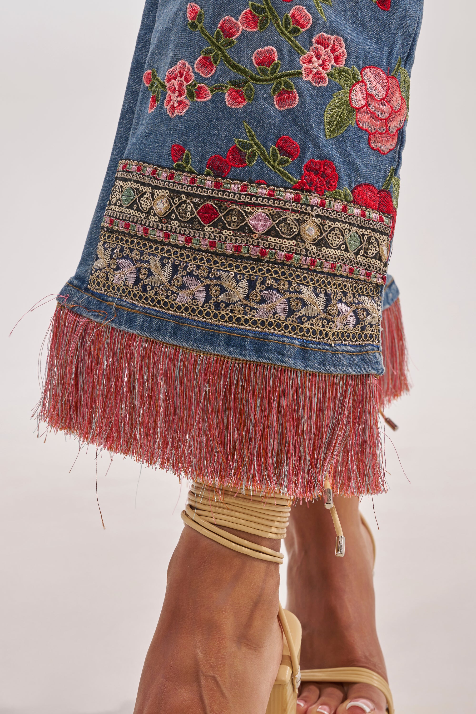 Close-up of a lower leg wearing FLORAL FOR DAYS STRAIGHT LEG DENIM featuring red floral embroidery, ornate trim, and red fringe at the hem, paired with beige strappy sandals. 10