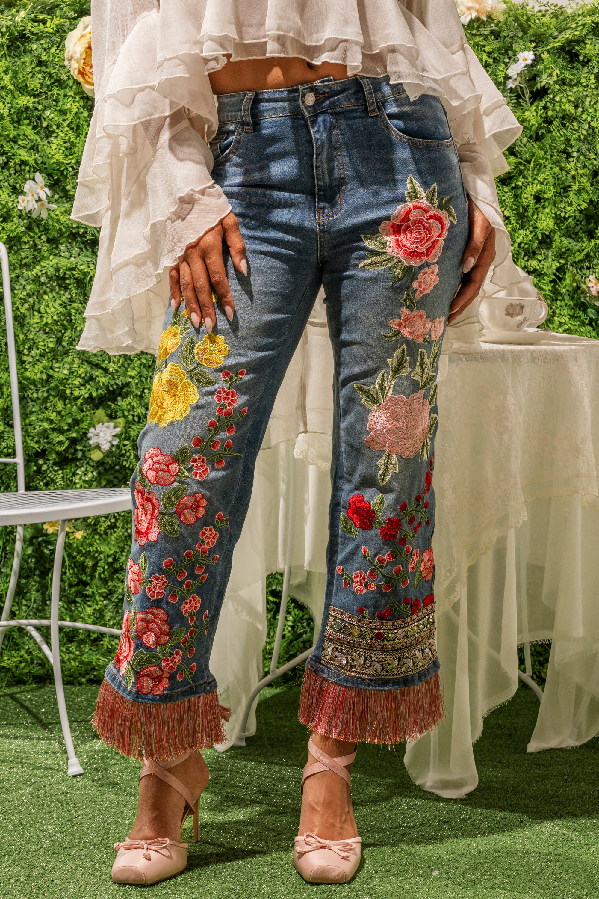 A person wears the FLORAL FOR DAYS STRAIGHT LEG DENIM featuring colorful floral embroidery and fringe, styled with a white ruffled blouse and pink satin heels, while standing on grass before a vibrant floral backdrop. 0