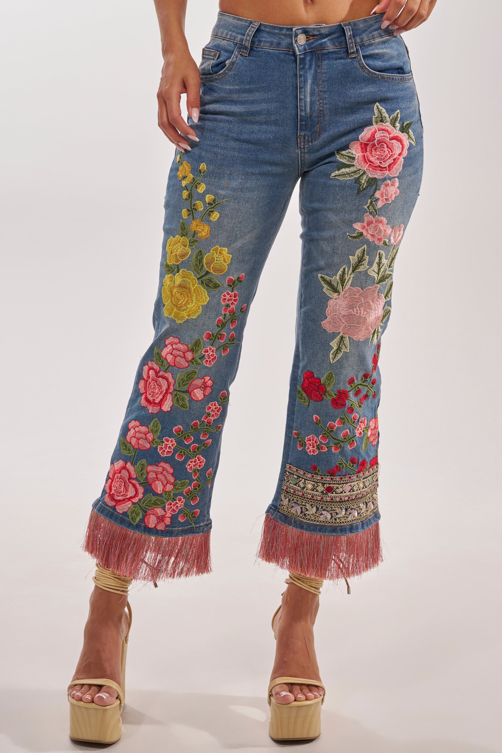 A woman wears the FLORAL FOR DAYS STRAIGHT LEG DENIM, featuring boho-luxe floral embroidery with colorful flowers and pink fringe at the hem, paired with tan platform sandals. 2