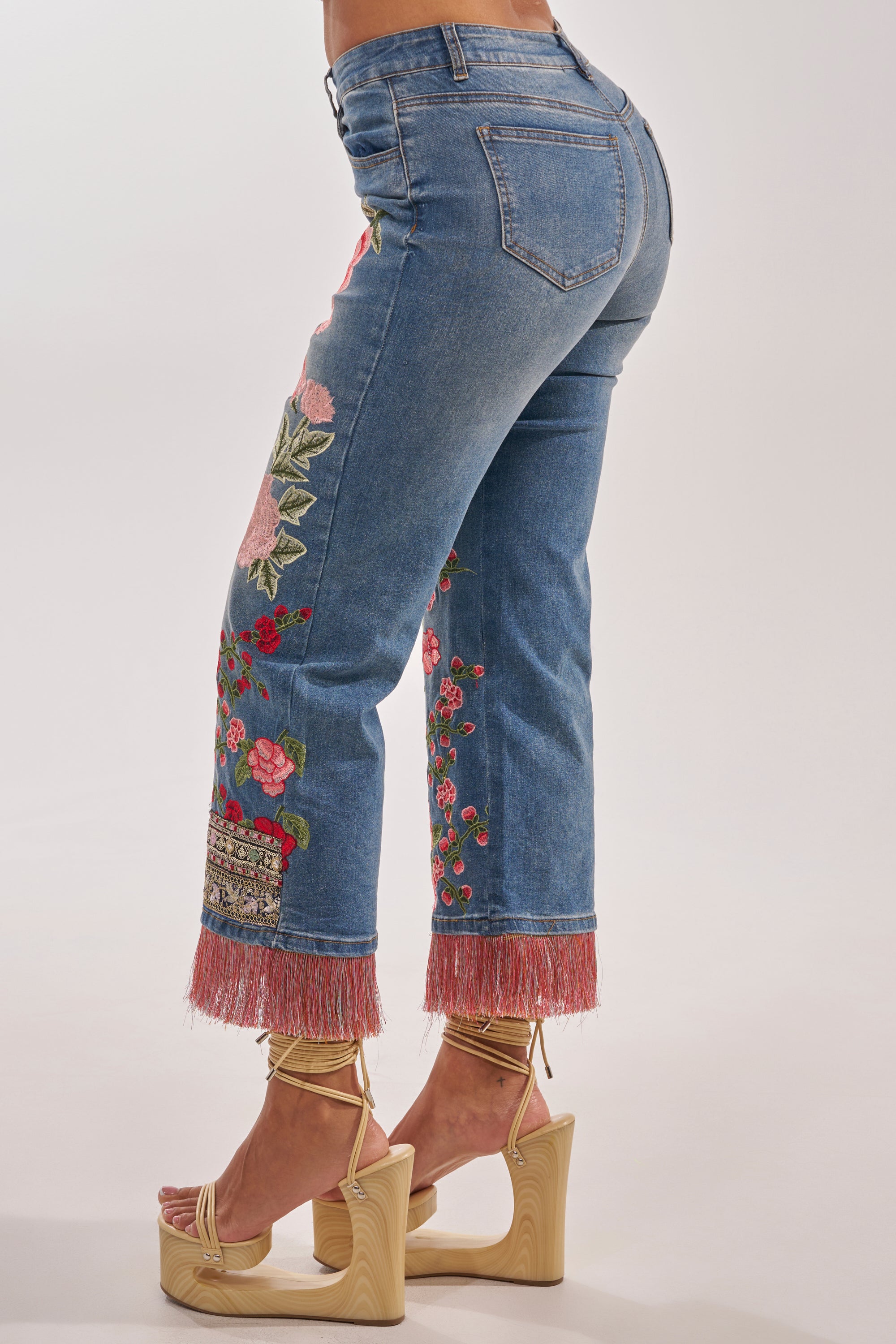 Wearing the FLORAL FOR DAYS STRAIGHT LEG DENIM with boho-luxe floral embroidery and pink fringe hems, paired with gold strappy platform heels, a person stands against a plain background. 3