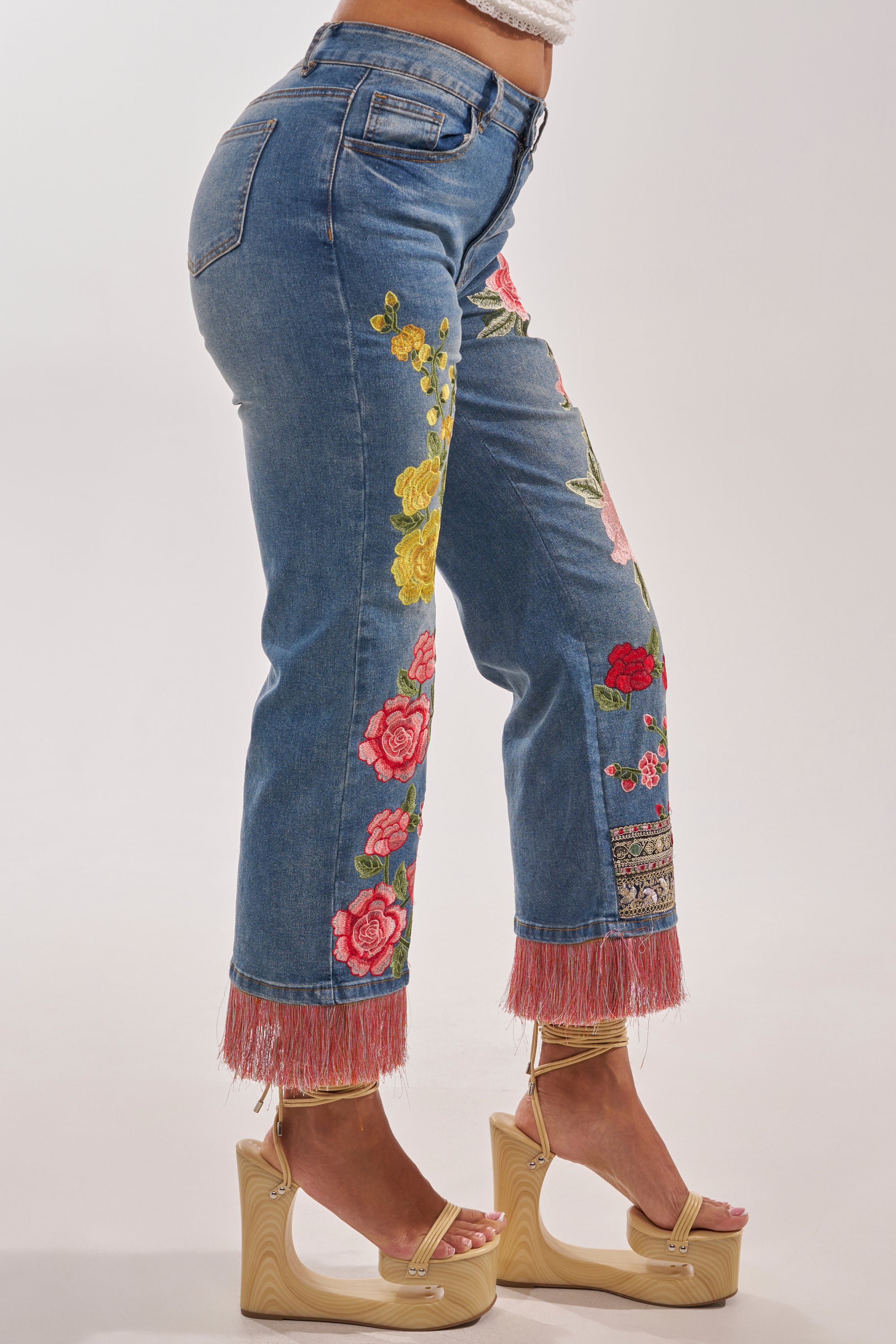 Someone wears the FLORAL FOR DAYS STRAIGHT LEG DENIM—boho-luxe jeans with colorful floral embroidery and pink fringe hem—paired with beige ankle-strap platform sandals, posing against a plain backdrop. 4