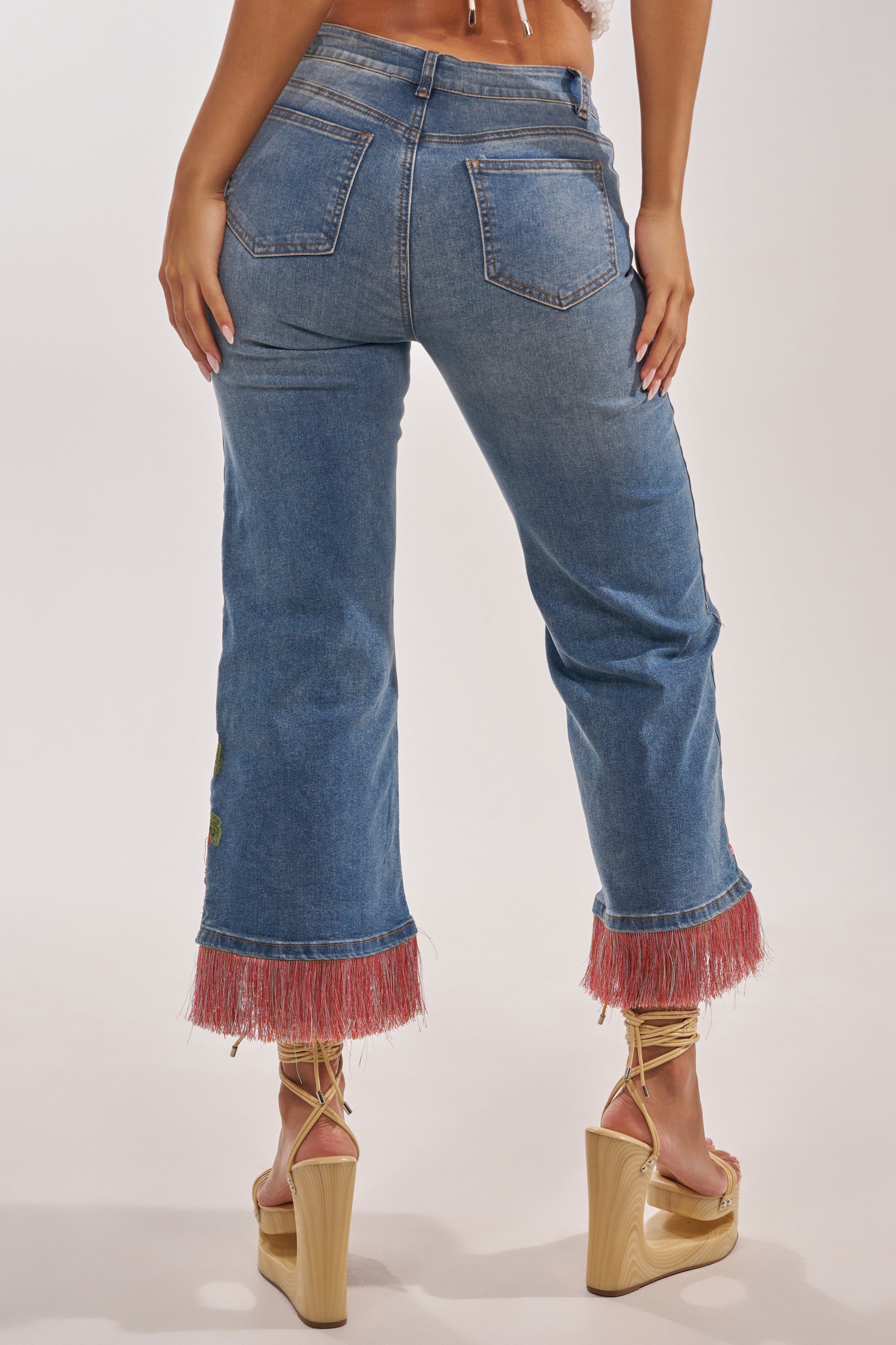 A person stands facing away, wearing FLORAL FOR DAYS STRAIGHT LEG DENIM with pink fringe at the hems and beige platform heels, making the boho-luxe jeans pop against a plain, light background. 5