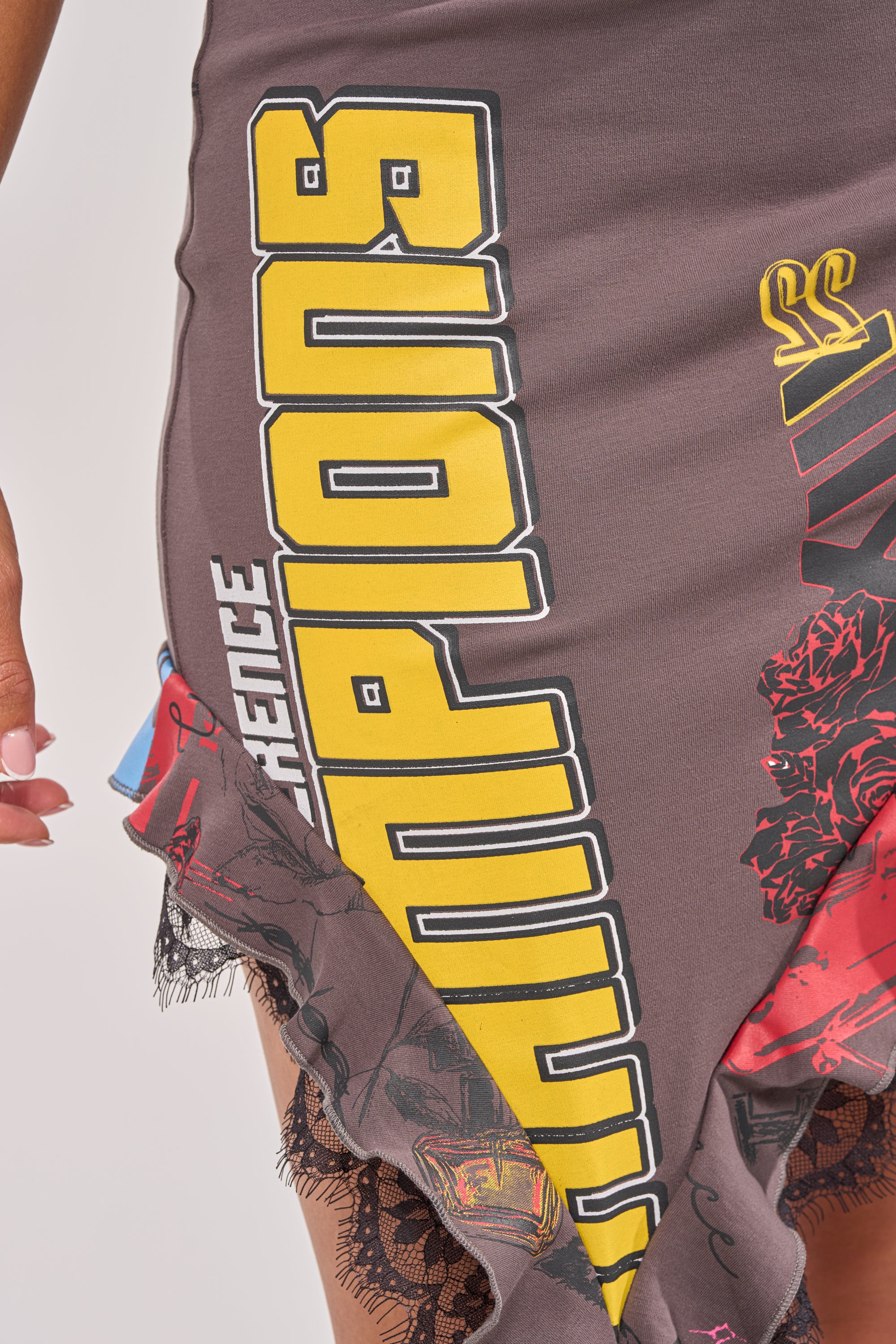 A close-up of a person wearing the BIG WINNER GRAPHIC SKIRT, featuring yellow "CHAMPIONS" text, multicolored graphics, lace trim, and red rose patterns. The person's left hand with light pink nail polish is visible. 7
