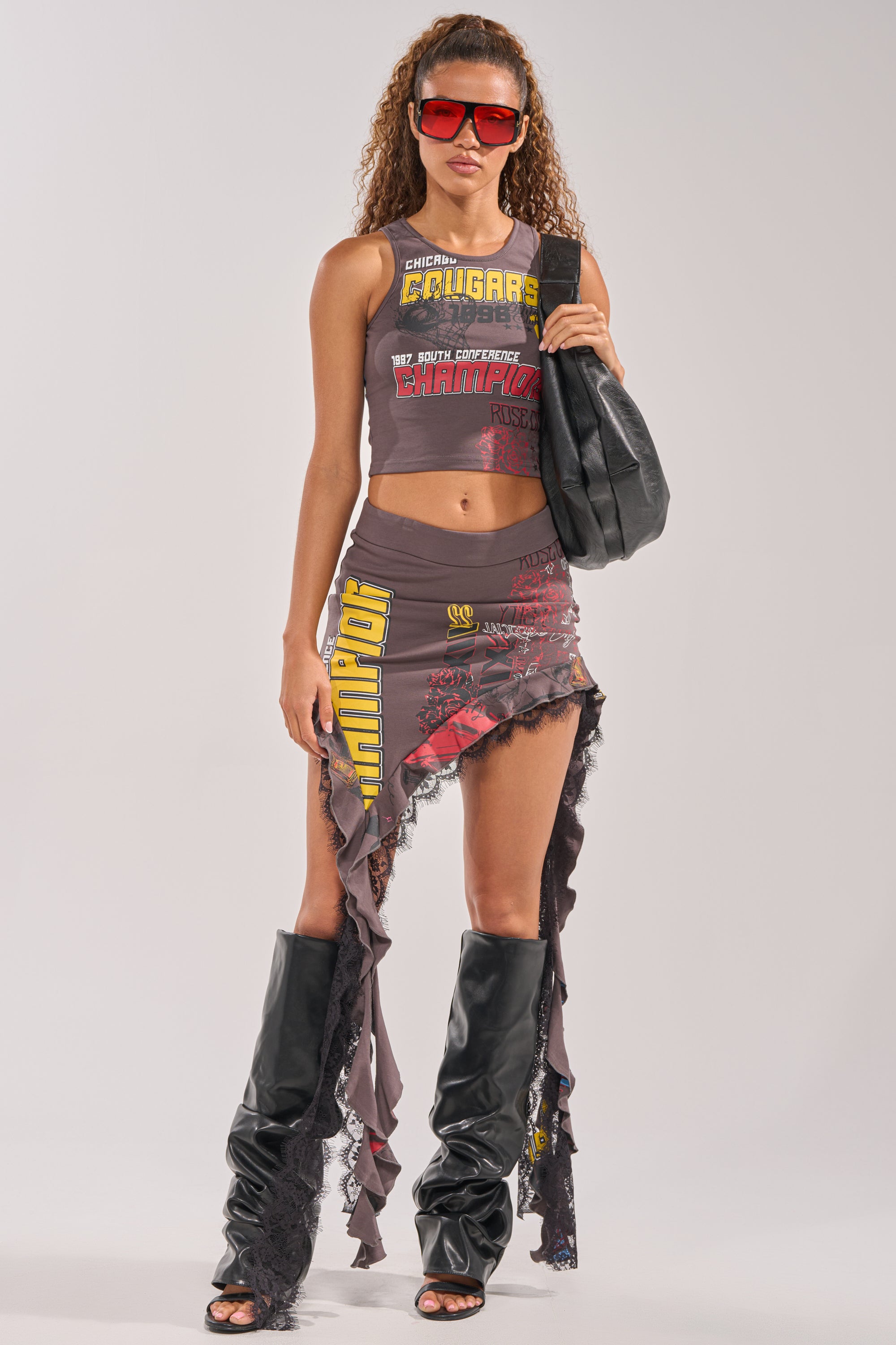 A woman with curly hair styles a BIG WINNER GRAPHIC TANK, red sunglasses, an asymmetrical text-ruffled skirt, lace-trimmed black slouchy boots, and a black bag against a plain backdrop for chic courtside flair. 5