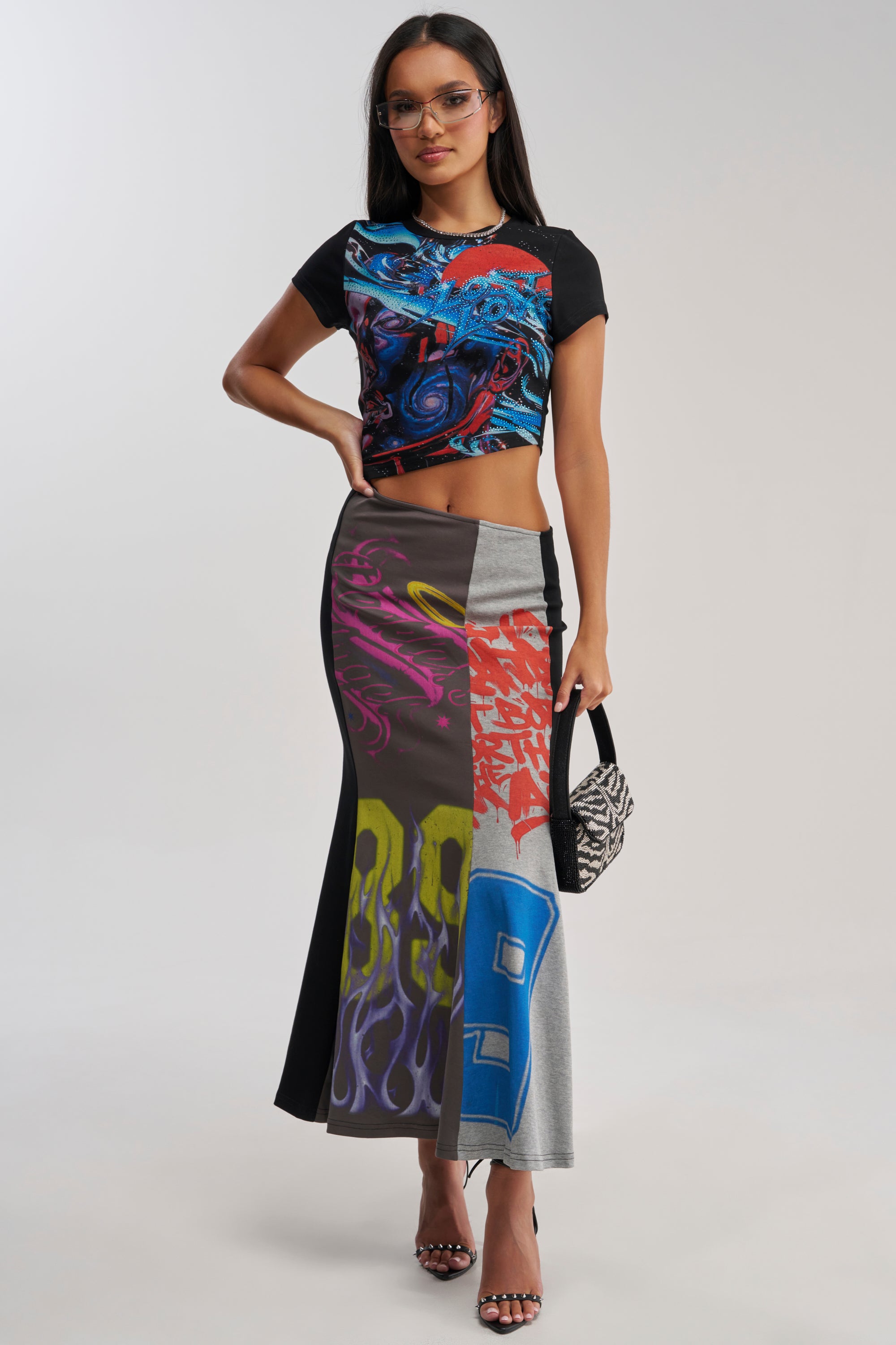 A woman wearing glasses, an abstract crop top, and the RACE YA GRAPHIC MAXI DRESS WITH SIDE CUTOUT stands confidently with one hand on her hip, holding a small black-and-white patterned handbag.