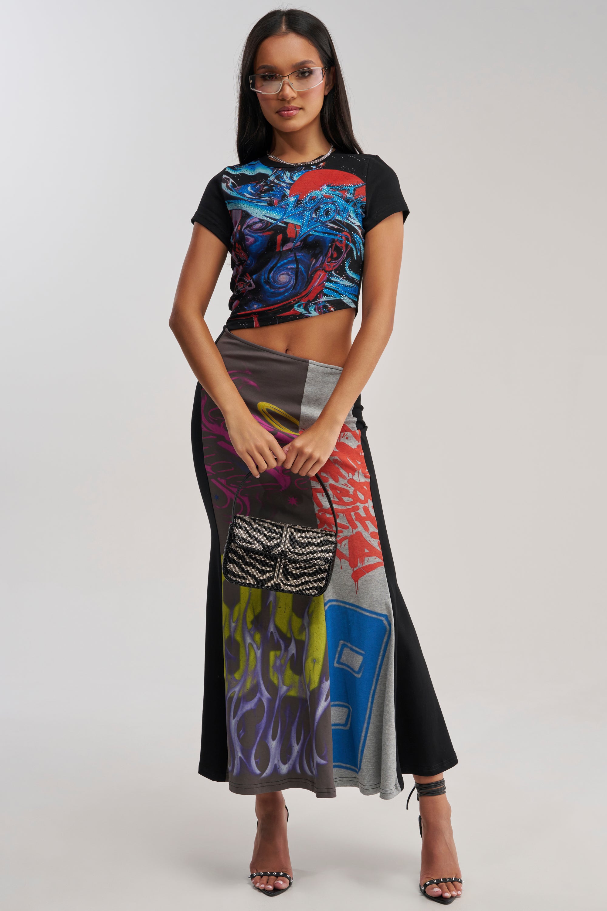 A woman in glasses wears the RACE YA GRAPHIC MAXI DRESS WITH SIDE CUTOUT and holds a small black-and-white animal print purse, standing confidently and looking at the camera.