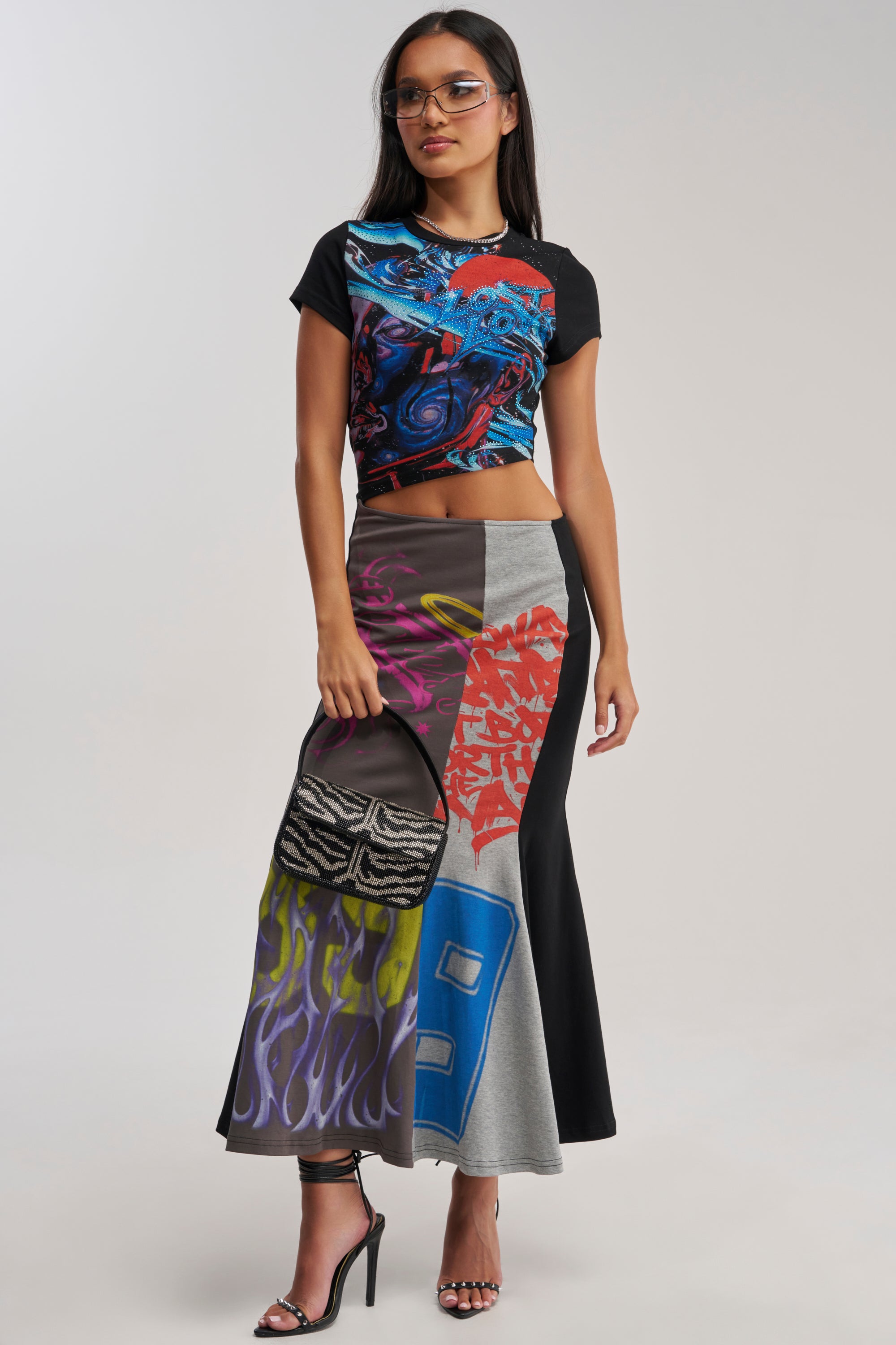 A woman with long, dark hair and glasses stands in heels, holding a small patterned handbag. She wears the RACE YA GRAPHIC MAXI DRESS WITH SIDE CUTOUT—a colorful statement streetwear piece.