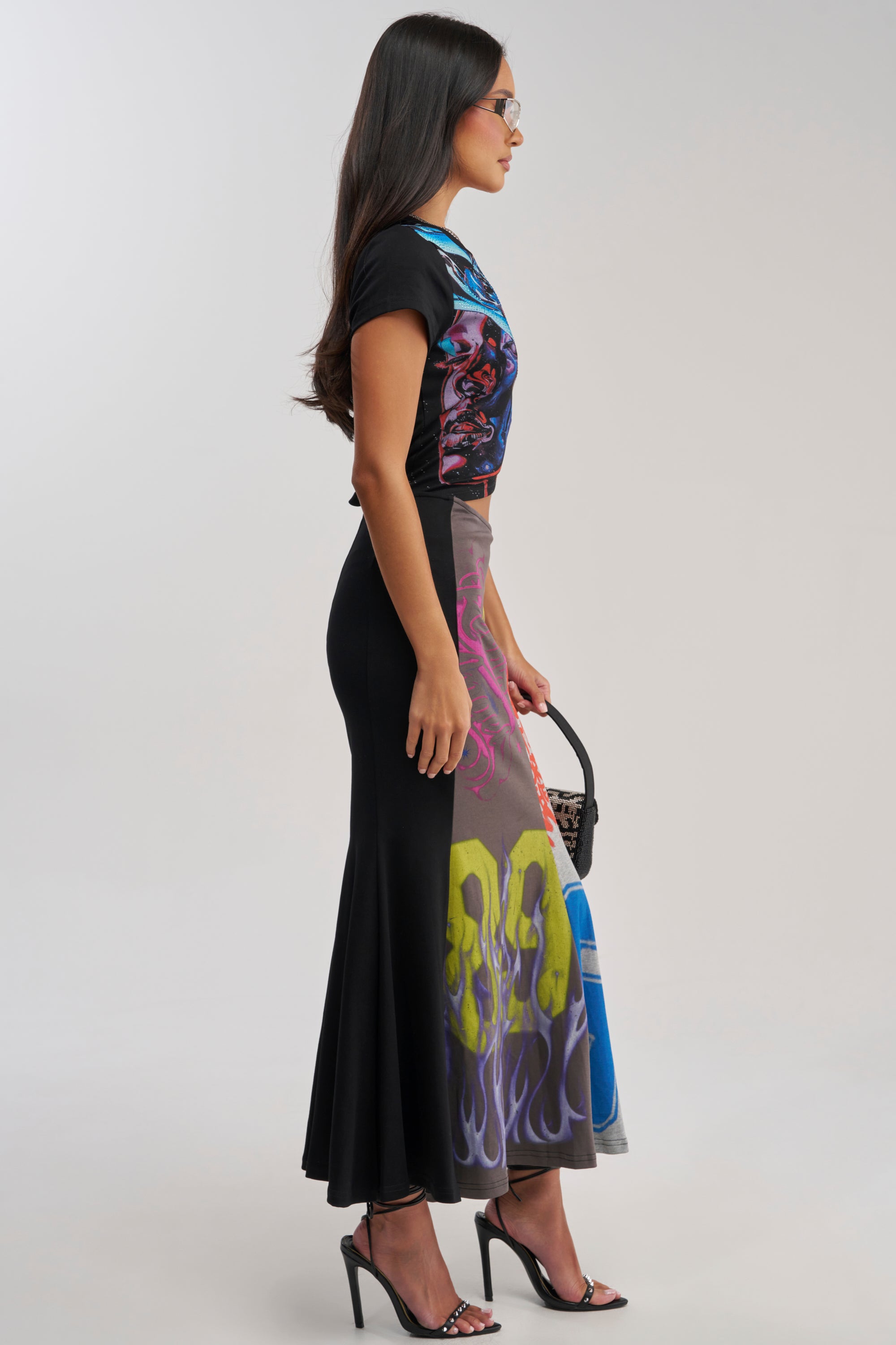 A woman with long dark hair and glasses stands in profile wearing the RACE YA GRAPHIC MAXI DRESS WITH SIDE CUTOUT, black heeled sandals, and holding a small black handbag.