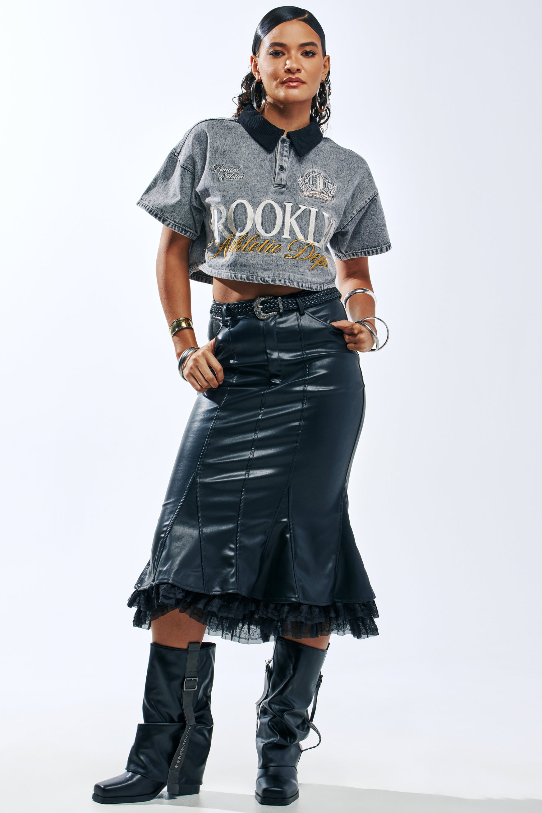 A person wears a black faux leather FEEL LIKE A QUEEN MIDI SKIRT with a ruffled hem, paired with a gray cropped polo shirt, a braided belt, and black calf-height boots.