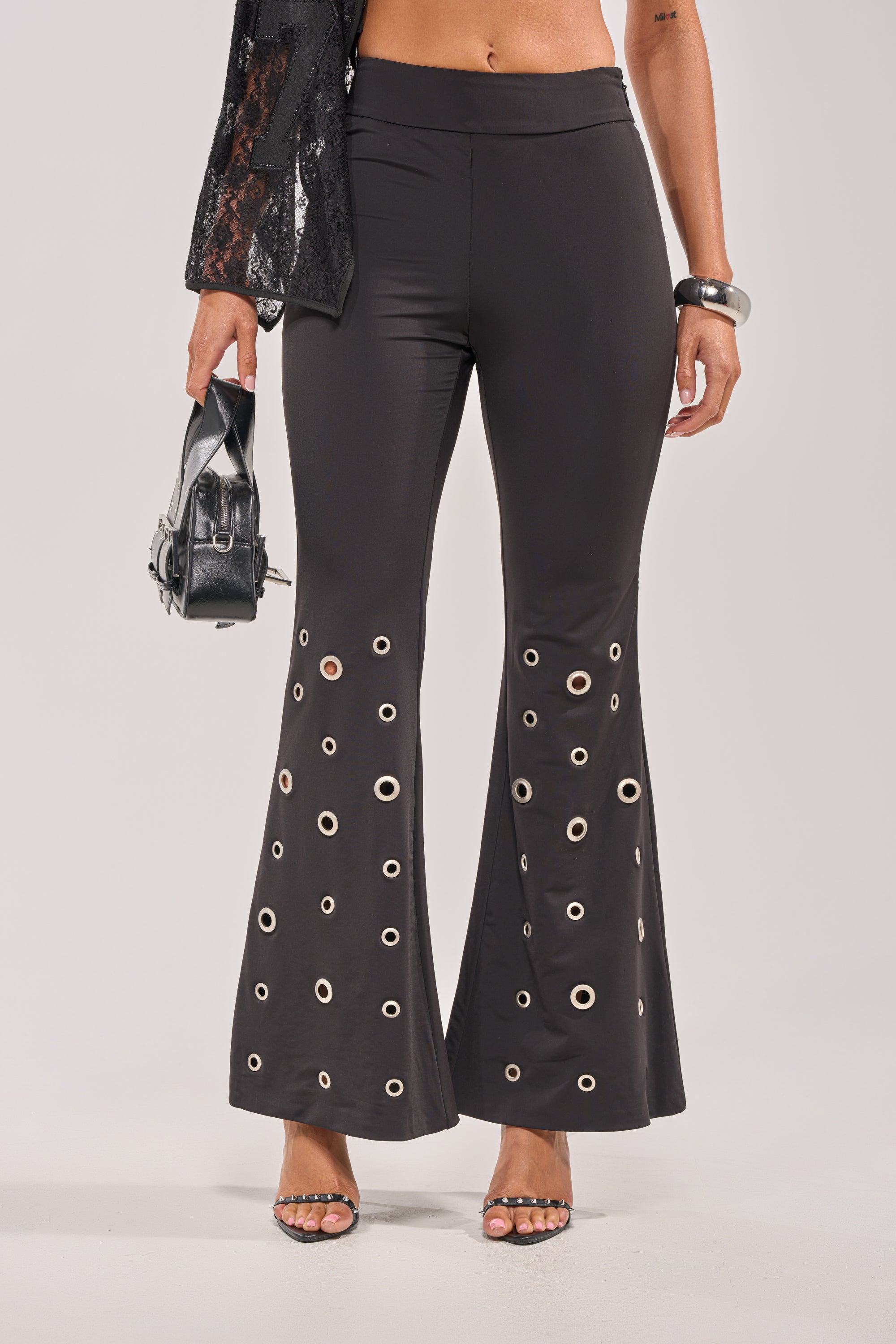 A person wears the CHECK IT OUT GROMMET FLARE LEGGING—black, high-rise with a silver grommet hem—styled with a small black bag, silver bangle, and open-toe heels. 0
