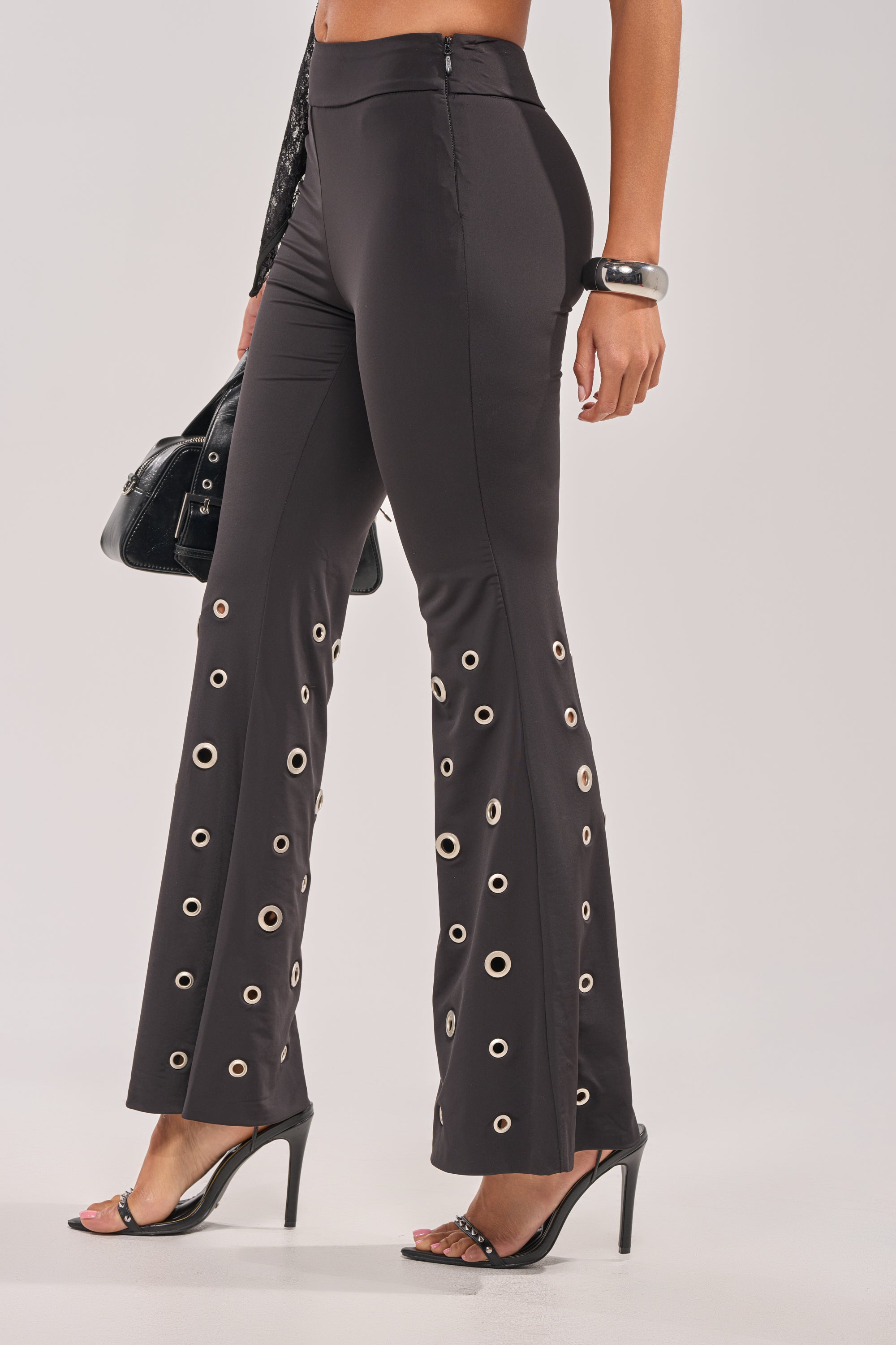 Wearing the CHECK IT OUT GROMMET FLARE LEGGING with a high-rise waist and silver grommet hem, she pairs it with a black sleeveless top, black heeled sandals, a chunky bracelet, and carries a sleek black handbag. 2