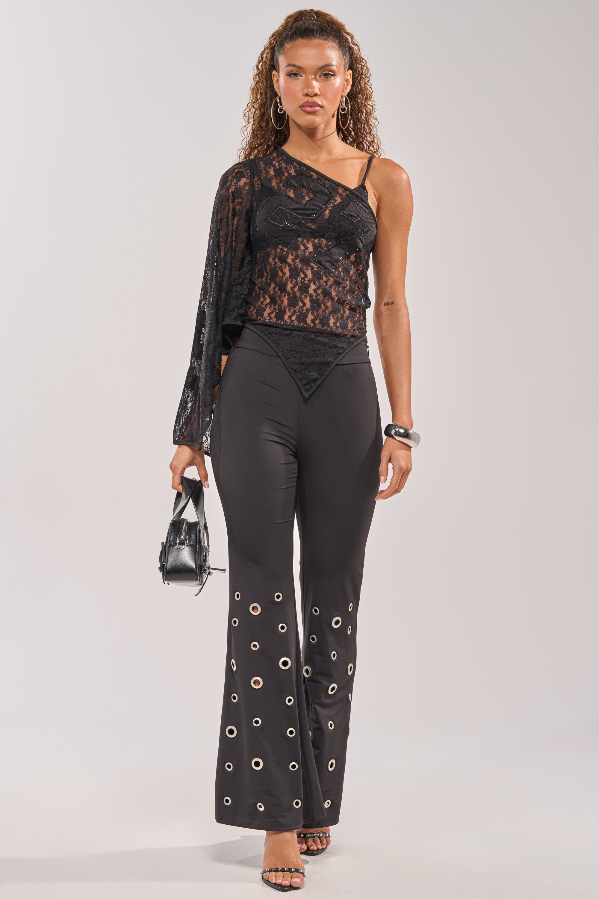 A woman models an asymmetrical black lace top, the CHECK IT OUT GROMMET FLARE LEGGING with a high-rise waistband and silver grommet hem, black heeled sandals, hoop earrings, a bangle, and carries a small black handbag. 5