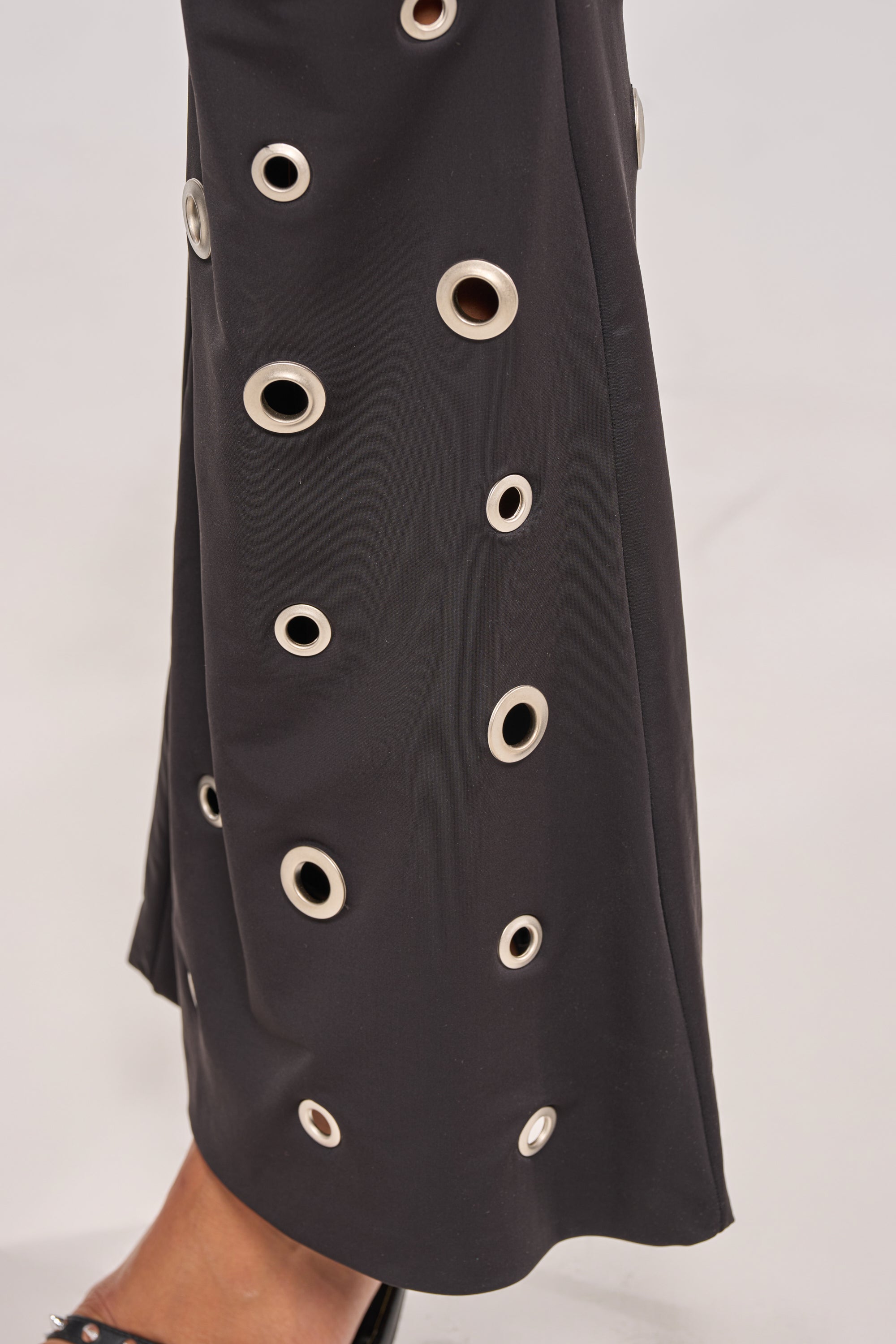 A close-up of the CHECK IT OUT GROMMET FLARE LEGGING in black shows multiple large and small silver grommets arranged irregularly; the lower leg appears at bottom left, emphasizing the eye-catching silver grommet hem. 7