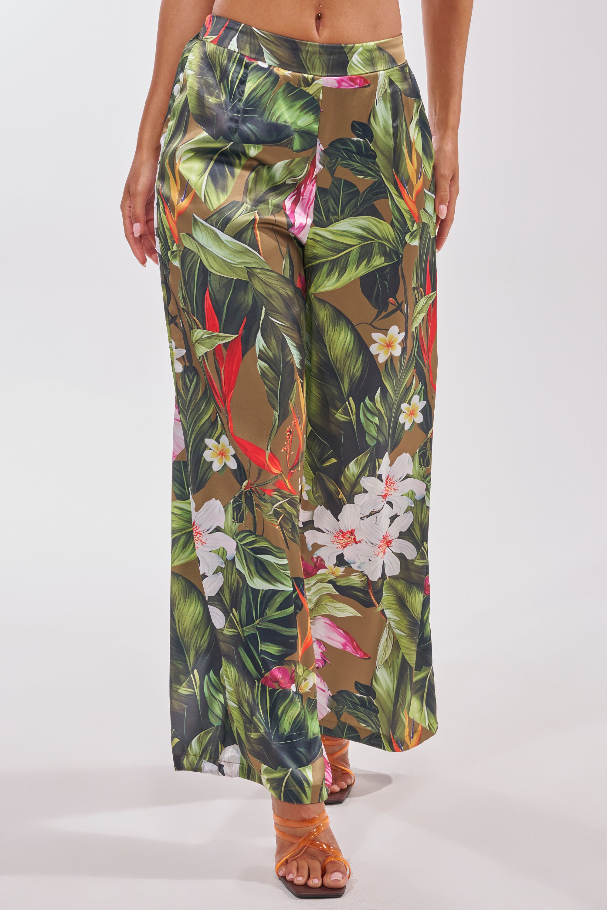 Model wears INTO THE WILD FLORAL TROUSER—high-waisted, wide-leg trousers with a green, red, and white tropical floral print—paired with strappy tan sandals. Perfect summer pants shown against a plain white background. 0
