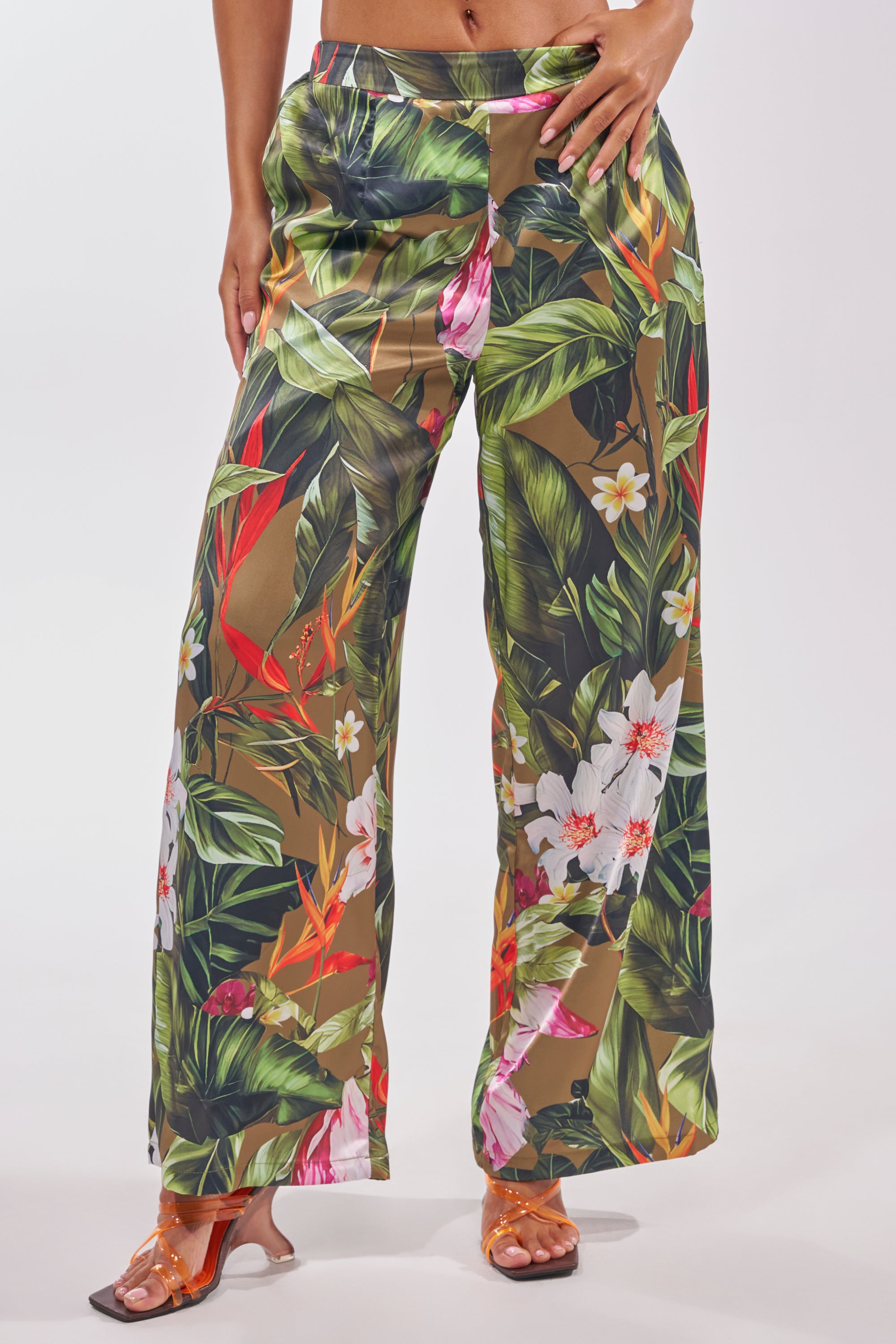 Woman wears the INTO THE WILD FLORAL TROUSER—high-waisted, wide-leg pants with a vibrant green, red, and white tropical floral print—paired with orange open-toe heels for a bold summer look. 1