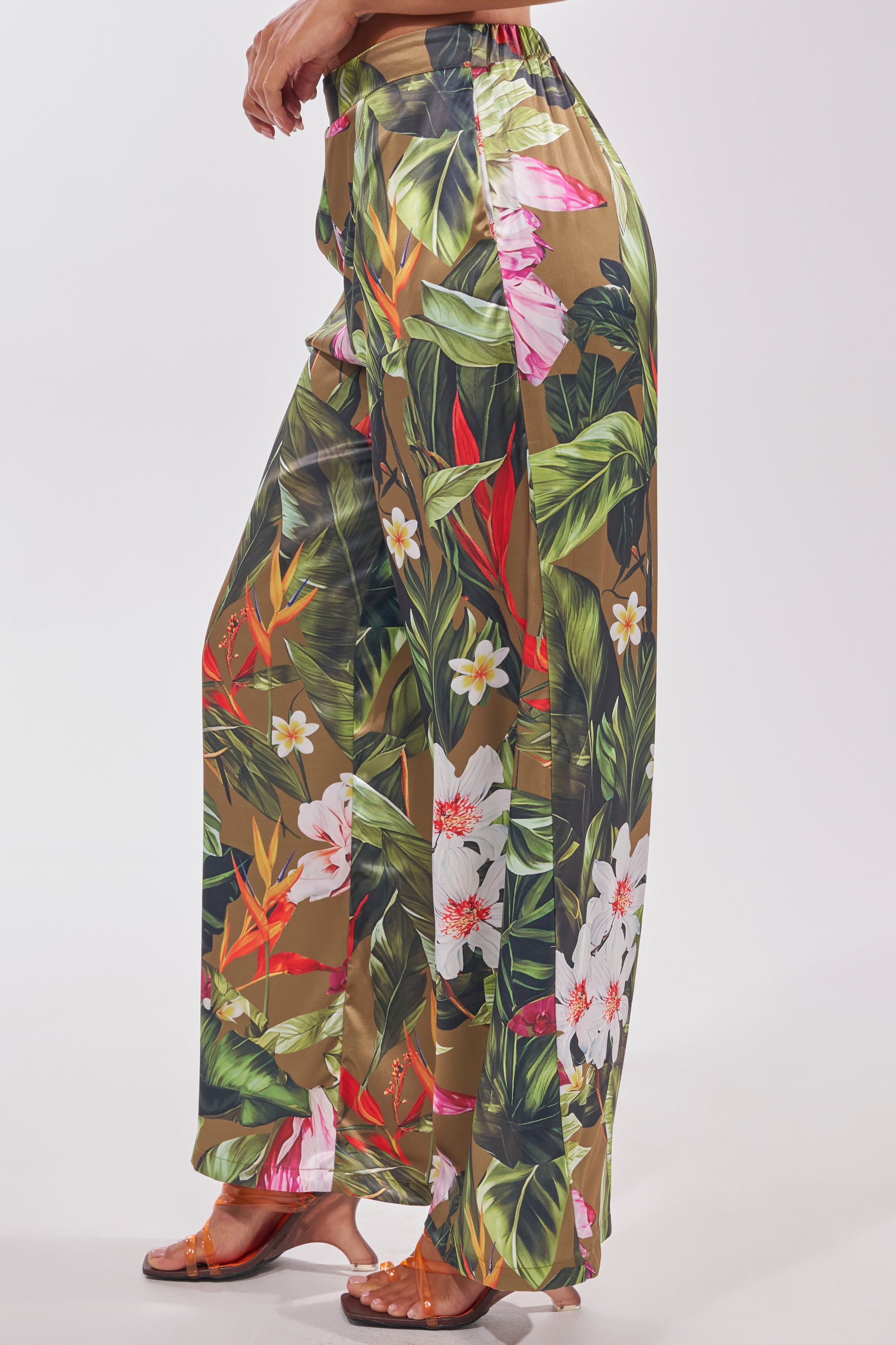 A person styles the INTO THE WILD FLORAL TROUSER—wide-leg satin pants with a vibrant tropical print—in green, white, pink, and orange, paired with flat brown sandals. The look is set against a plain light background. 2