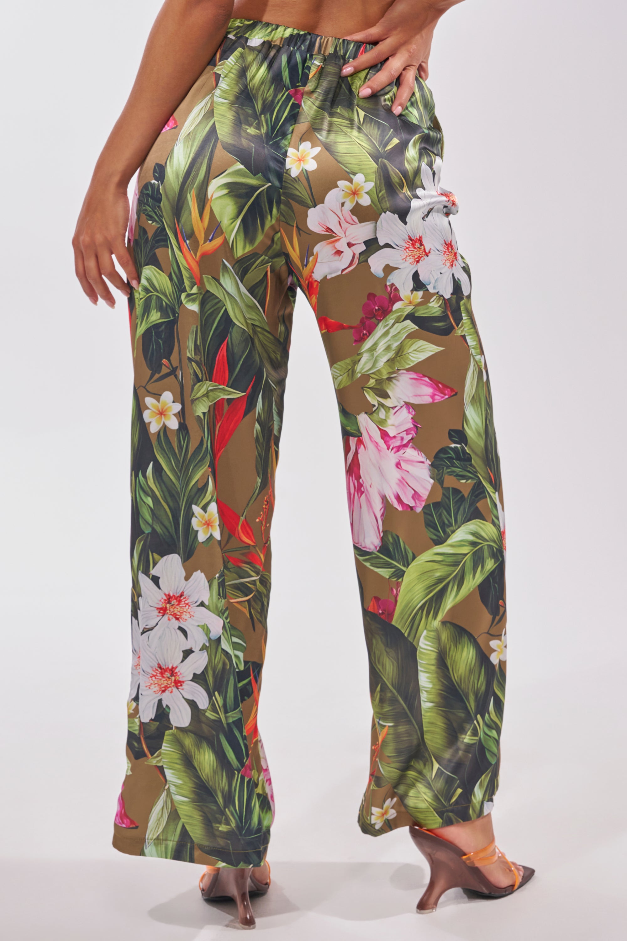 Seen from the back, a person wears the INTO THE WILD FLORAL TROUSER—tropical print satin pants with vibrant floral and leaf motifs—styled with heeled sandals for chic, effortless summer style. 3