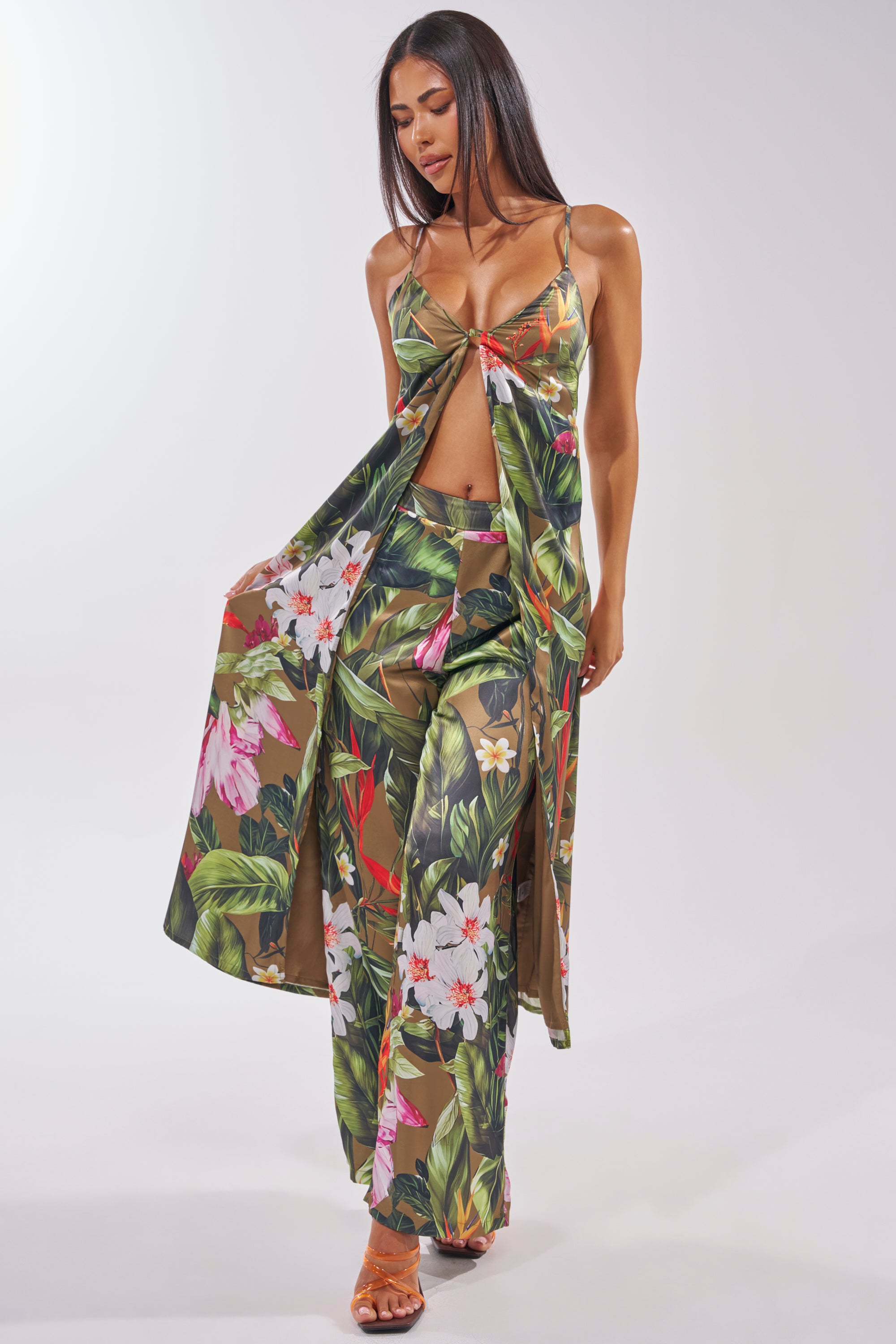 A woman poses against a light background in the INTO THE WILD FLORAL TROUSER set—tropical print satin with a long open-front top and matching wide-leg trousers—paired with strappy sandals, smiling gently as she looks down. 4