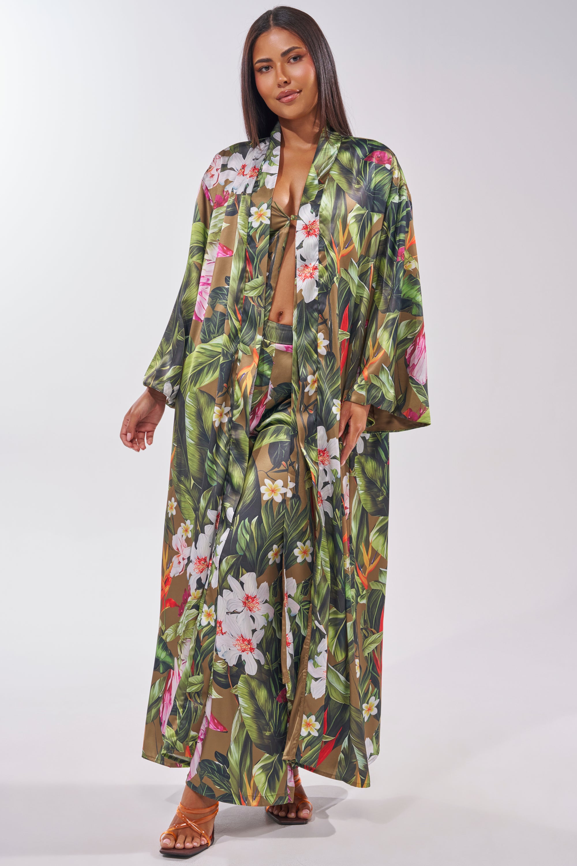A woman poses against a plain light background in the INTO THE WILD FLORAL TROUSER, styled with matching vacation wear and tan sandals. She has straight dark hair and completes the tropical floral look with a flowing sheer robe. 5