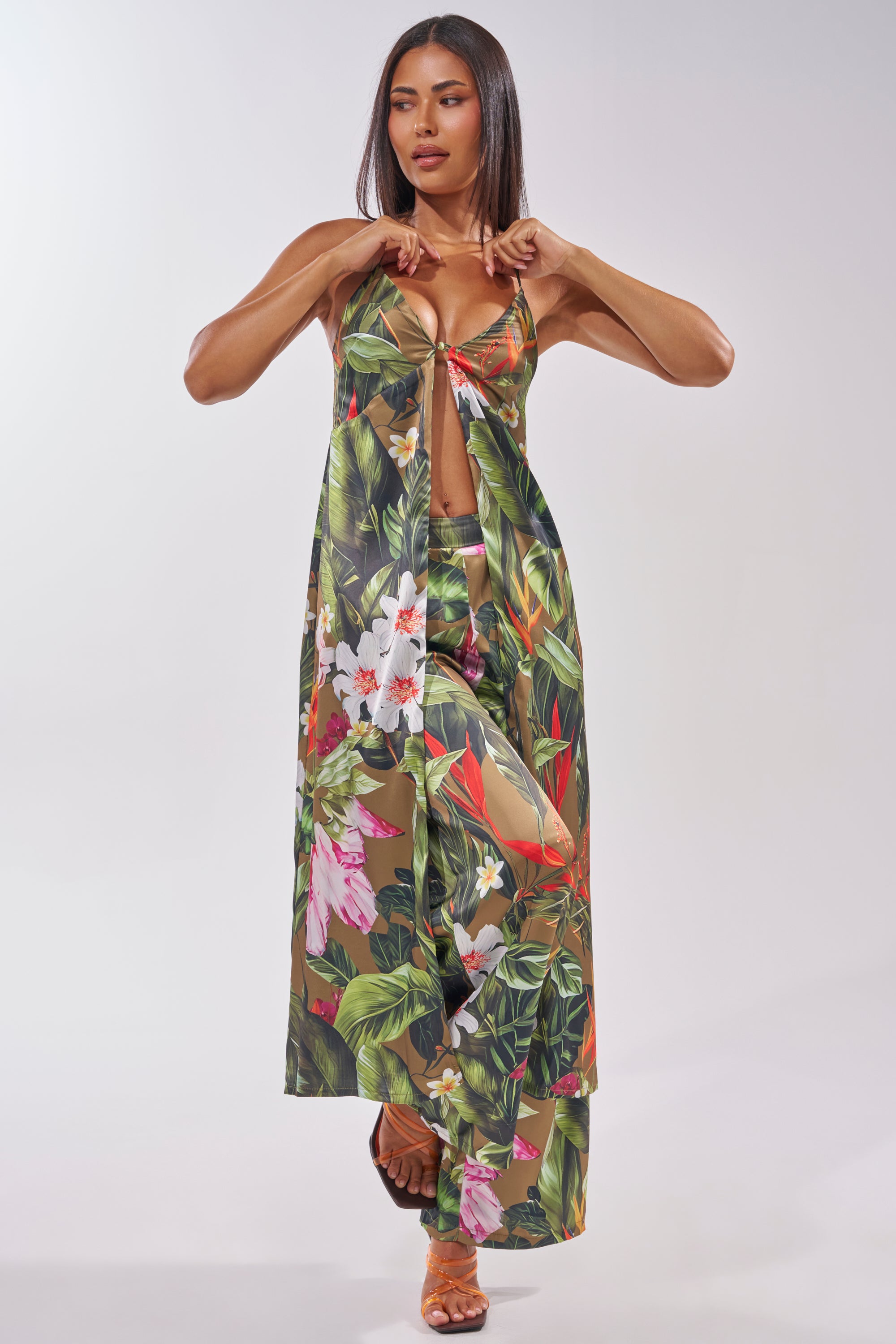 A woman adjusts the straps of a sleeveless tropical floral maxi dress with a front cutout, paired with heeled sandals and INTO THE WILD FLORAL TROUSER, set against a plain background. 7