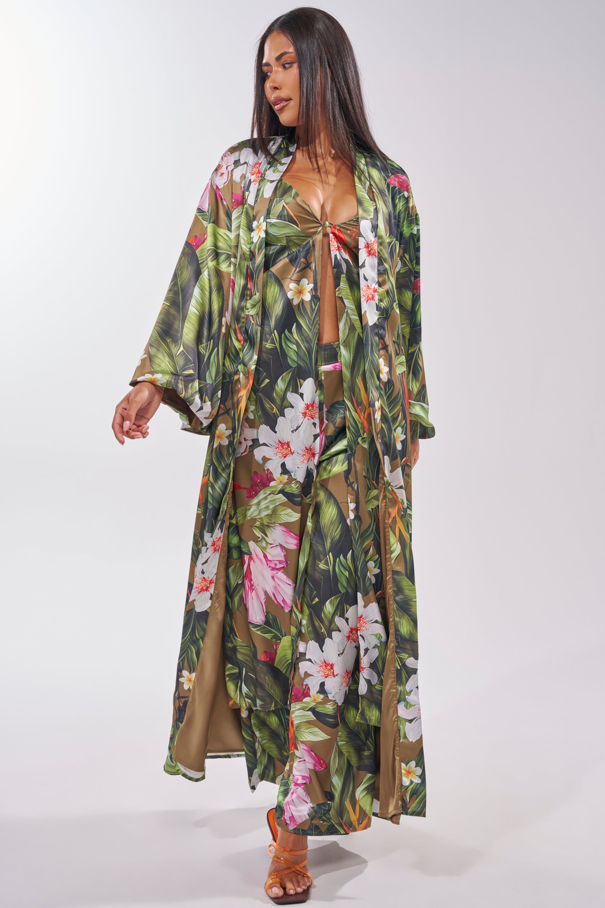 A woman stands against a light background, looking to the side, wearing the INTO THE WILD FLORAL TROUSER with a matching vibrant tropical print robe and orange heels. 8