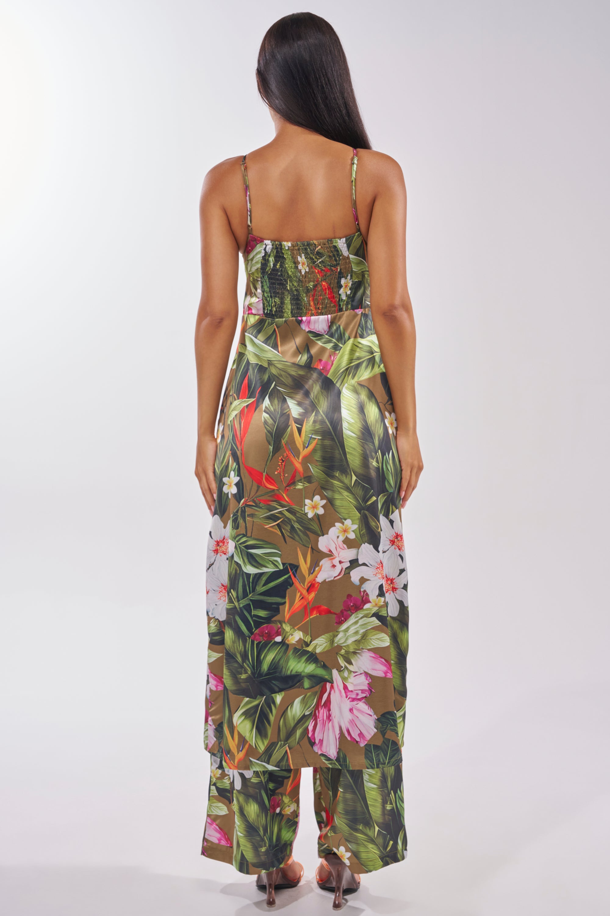 A woman with long dark hair, seen from behind, models the INTO THE WILD FLORAL BLOUSE—an ankle-length, sleeveless tropical print dress in vibrant green, red, and white—styled perfectly for summer against a plain light backdrop. 4