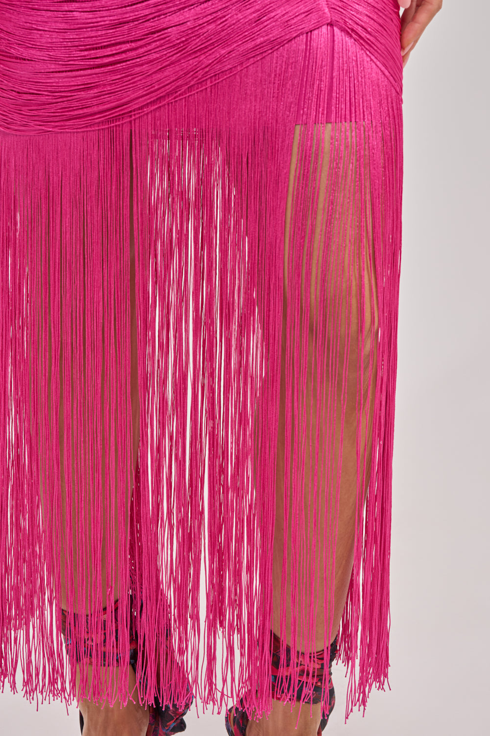 A close-up of a person in the TAKE ME DANCING FRINGE MAXI DRESS IN FUSCHIA, featuring long fuchsia fringe that partially reveals the legs. Set against a plain background, this vibrant maxi is perfect for making a statement at any special occasion. 7