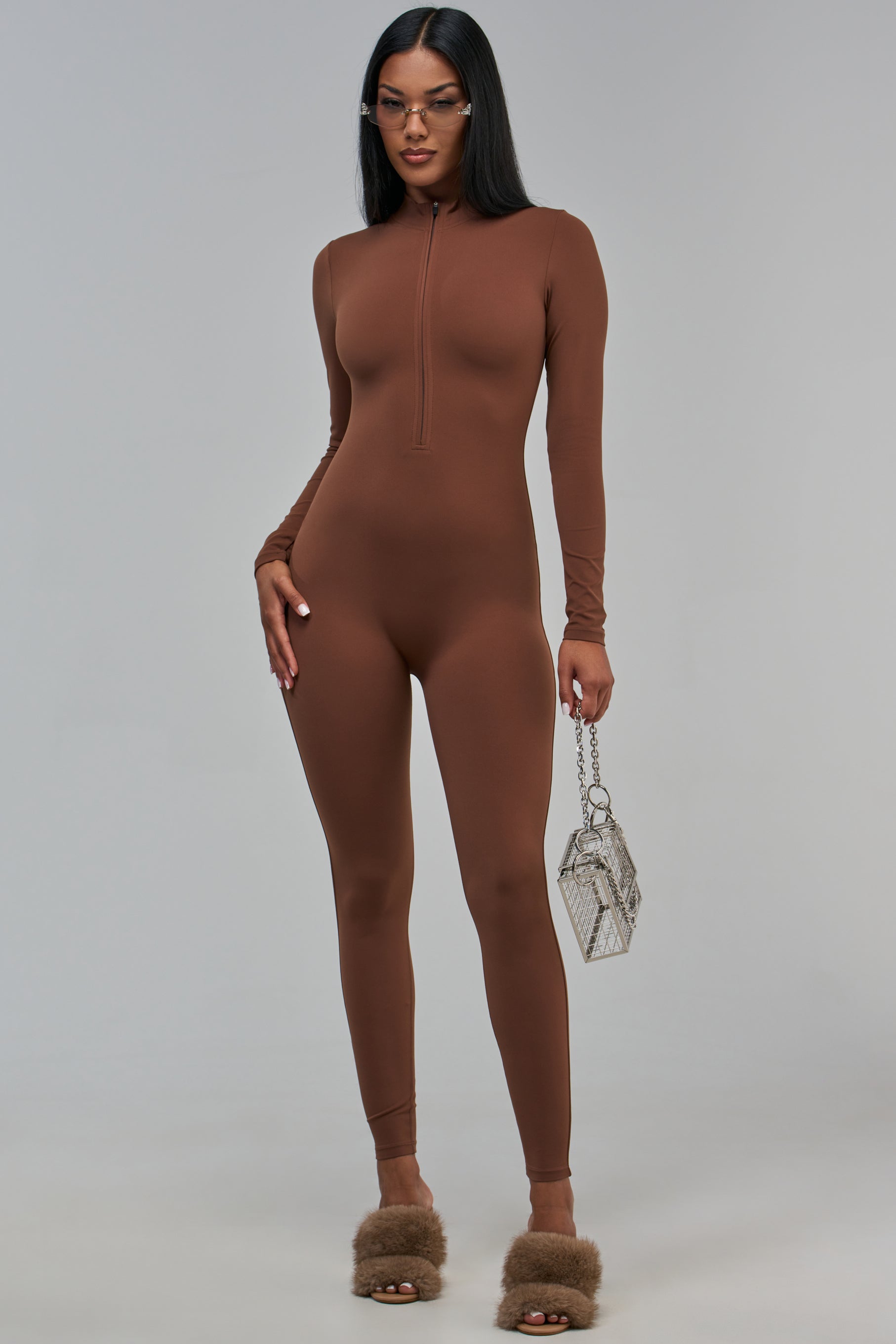 A woman wears a brown HOT MESS EXPRESS HIGH NECK ZIP-UP JUMPSUIT with fuzzy brown slides and a metallic boxy handbag.