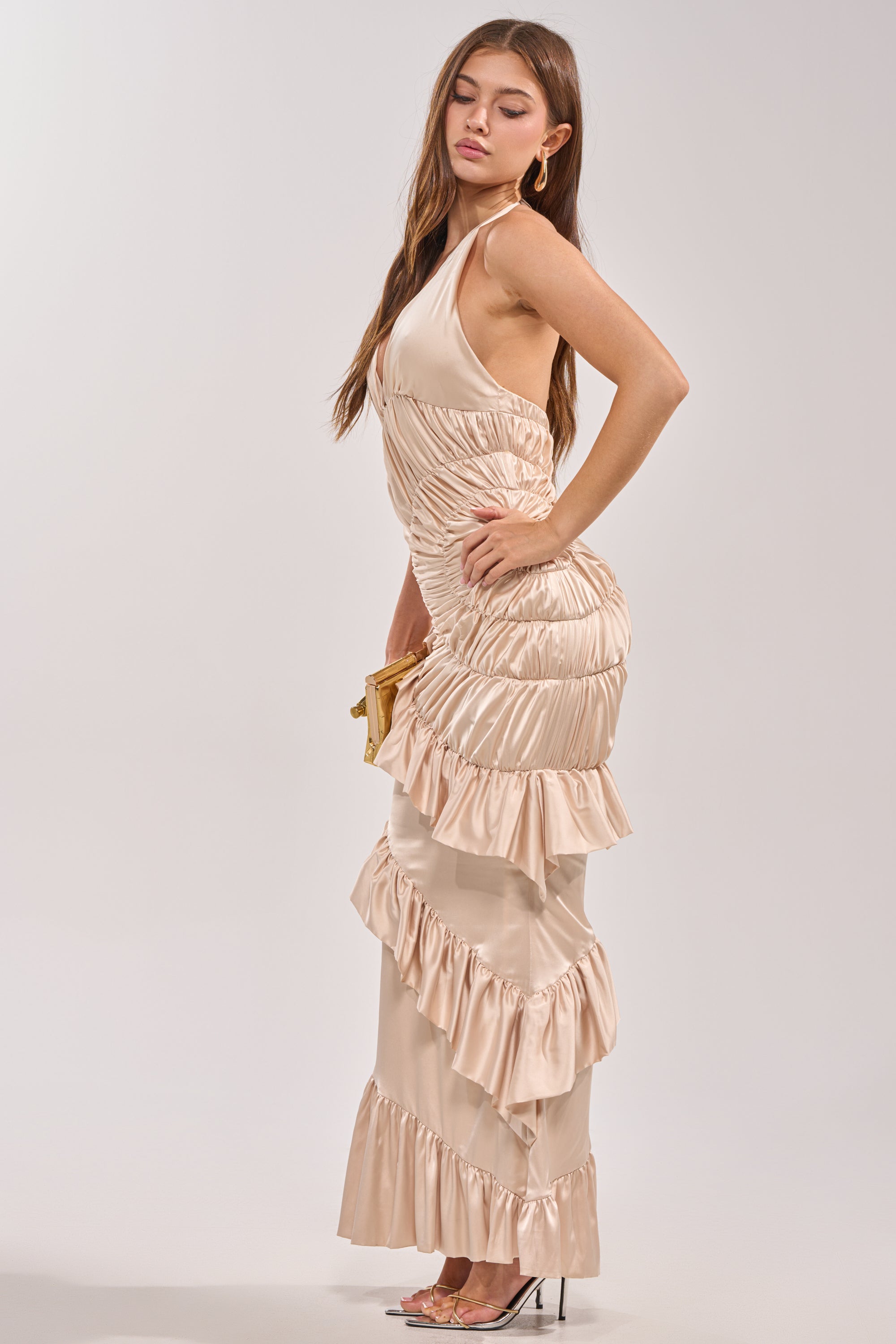 A woman models the MAKE YOU MINE RUFFLE SATIN MAXI DRESS—a shiny, cream ruched maxi with halter neck ruffles. She poses in profile with long brown hair, gold hoops, clear heels, and a gold clutch against a plain light gray background.