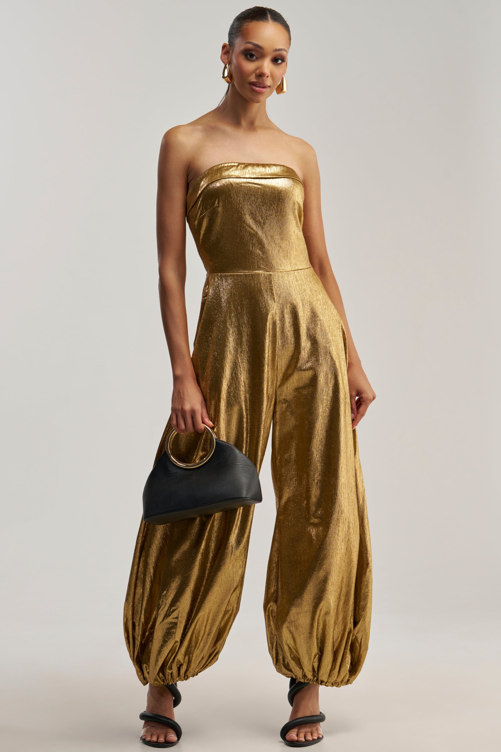 A woman models the MAKE IT LOUD METALLIC STRAPLESS JUMPSUIT, featuring a striking gold finish and balloon-style pants. She accessorizes with a black handbag and black heeled sandals while posing against a plain light background.