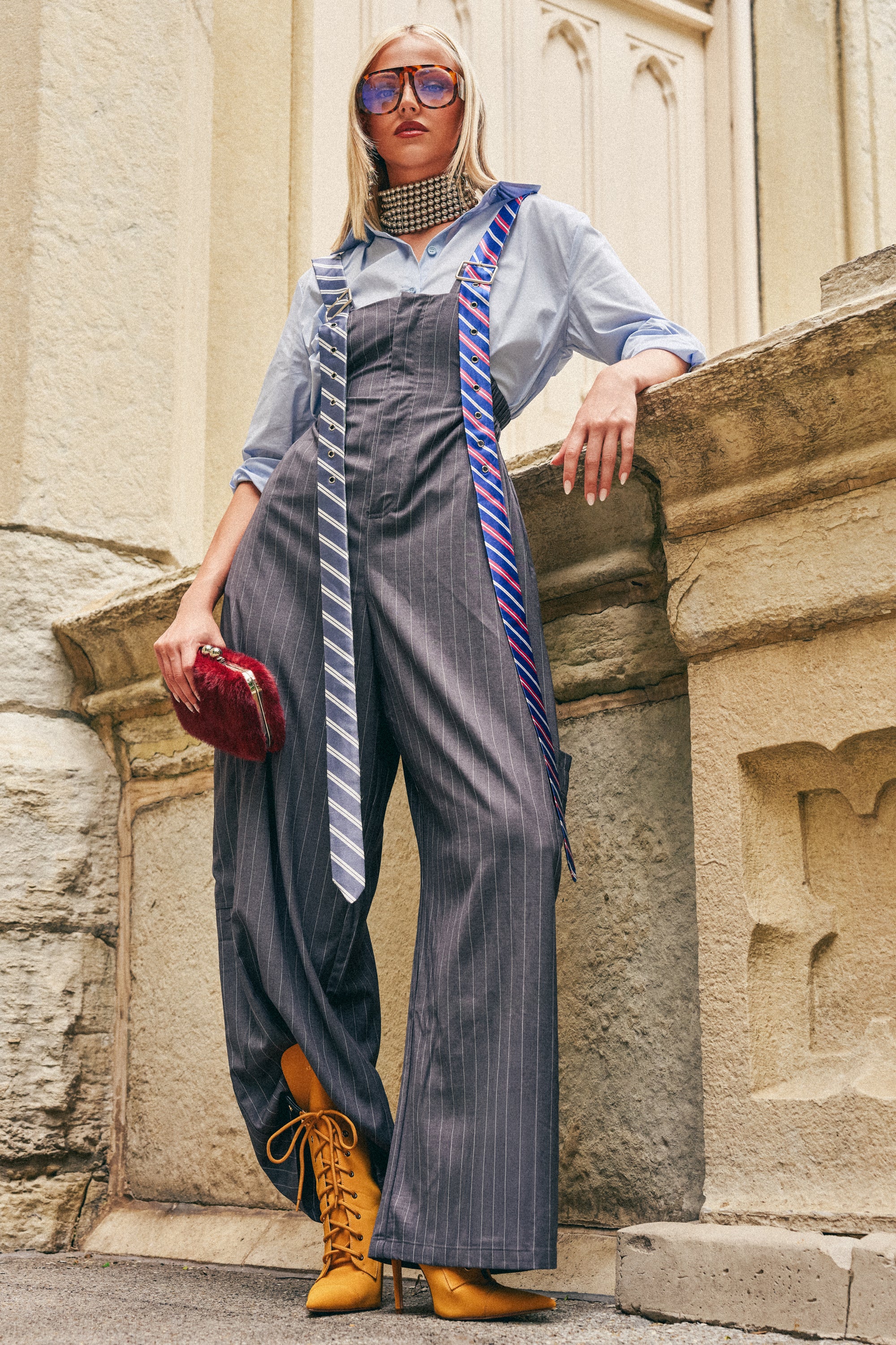 Wearing the CLASS ACT PINSTRIPE OVERALLS in grey, a woman poses confidently in a button-up shirt and yellow boots. She accessorizes with two neckties, sunglasses, a choker, and holds a red clutch against an ornate backdrop of light-colored stone architecture.