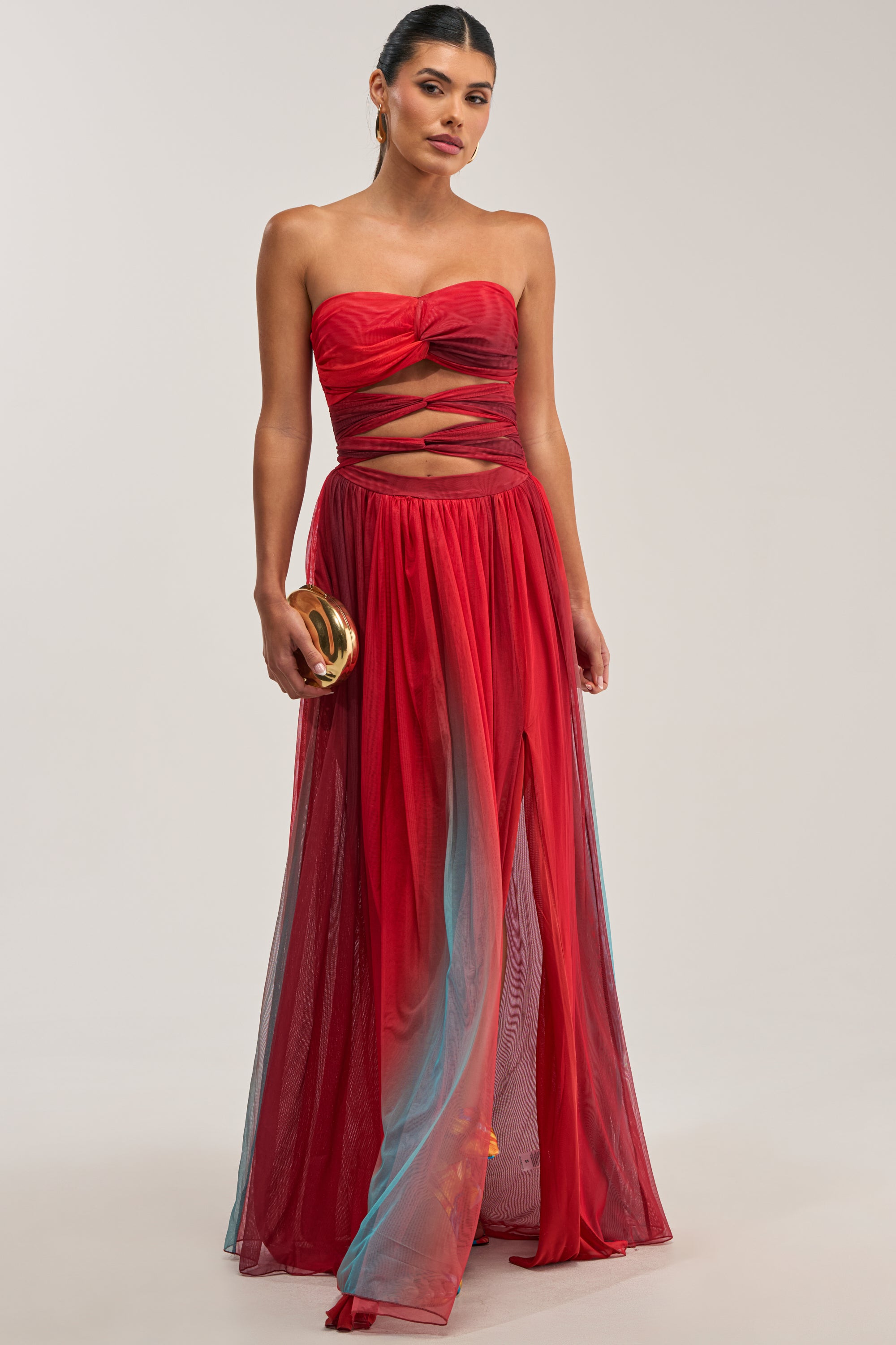 Woman wearing the CELESTE CHIFFON MAXI DRESS IN MULTI, featuring a strapless design with sheer bodice cutouts and a flowing chiffon evening skirt, holding a gold clutch and standing against a plain light background.