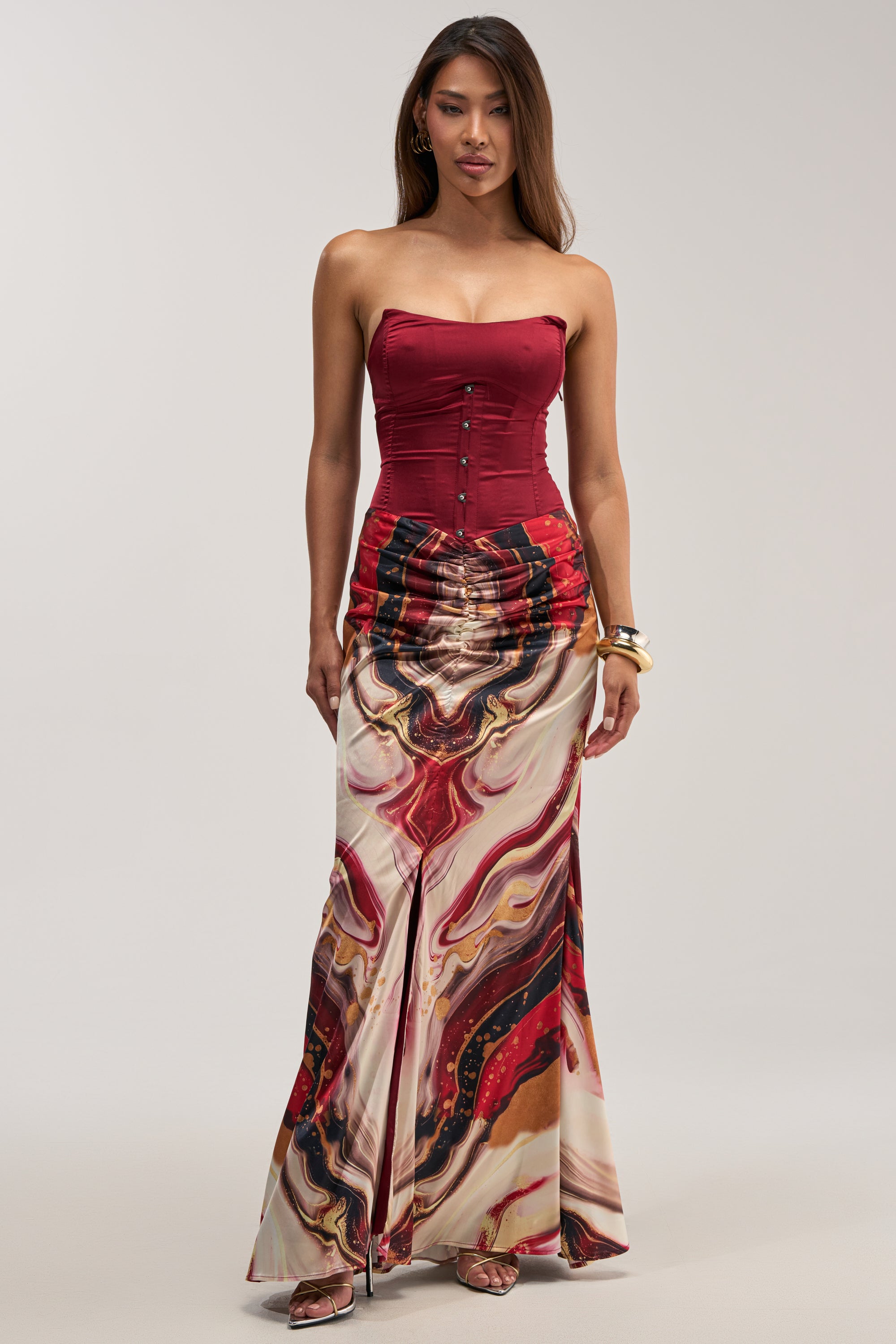 Dressed in the SAMARA ABSTRACT MAXI DRESS IN RED MULTI—a strapless maxi dress featuring a flowing skirt with a striking red, black, and cream marble pattern—a woman stands against a plain light background, making it the perfect elegant choice for a wedding guest.