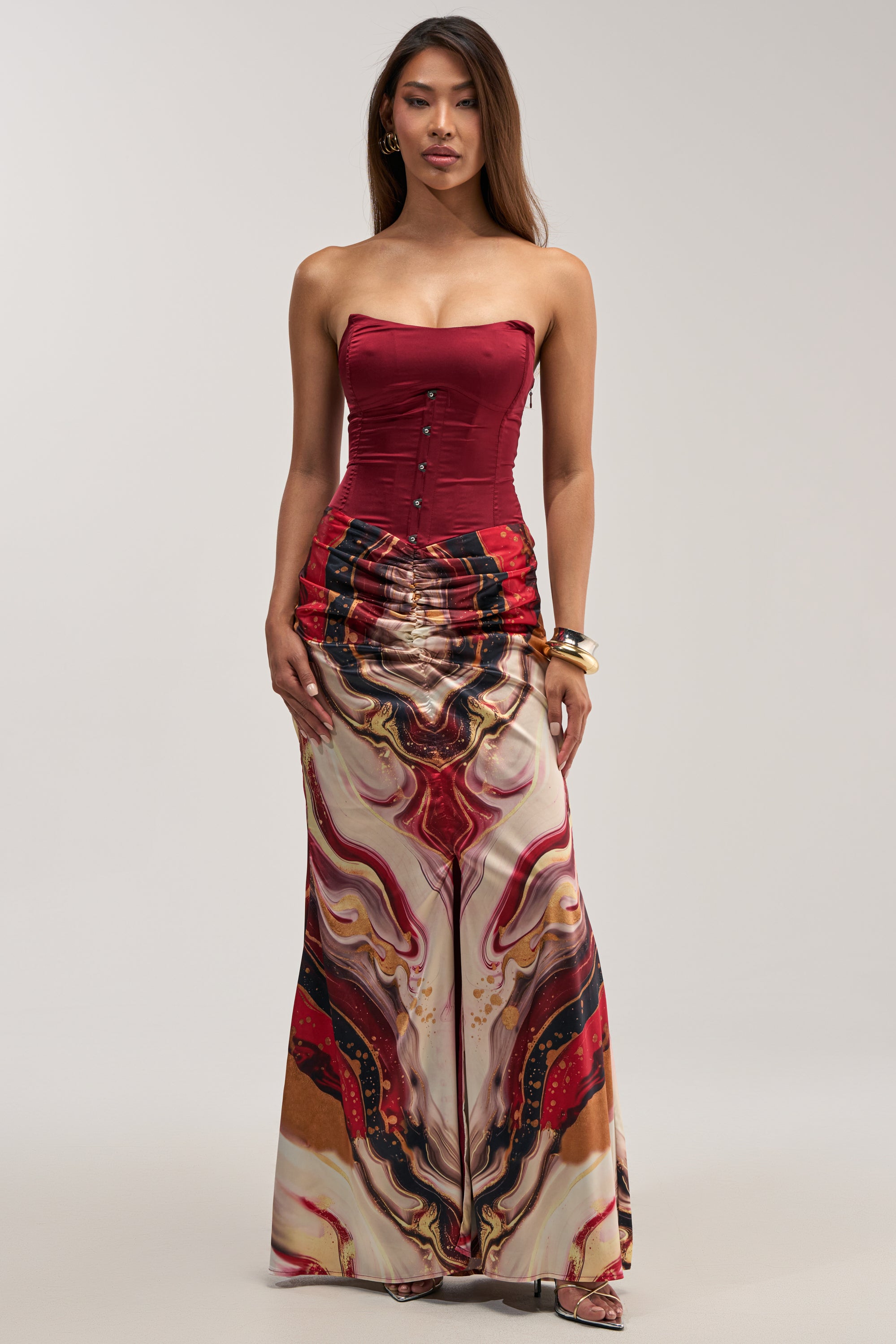 A woman stands against a plain light background wearing the SAMARA ABSTRACT MAXI DRESS IN RED MULTI—a strapless burgundy corset maxi dress with a long, high-waisted skirt adorned in a colorful, swirling abstract pattern. Perfect as a wedding guest dress, this look combines elegance with bold design.