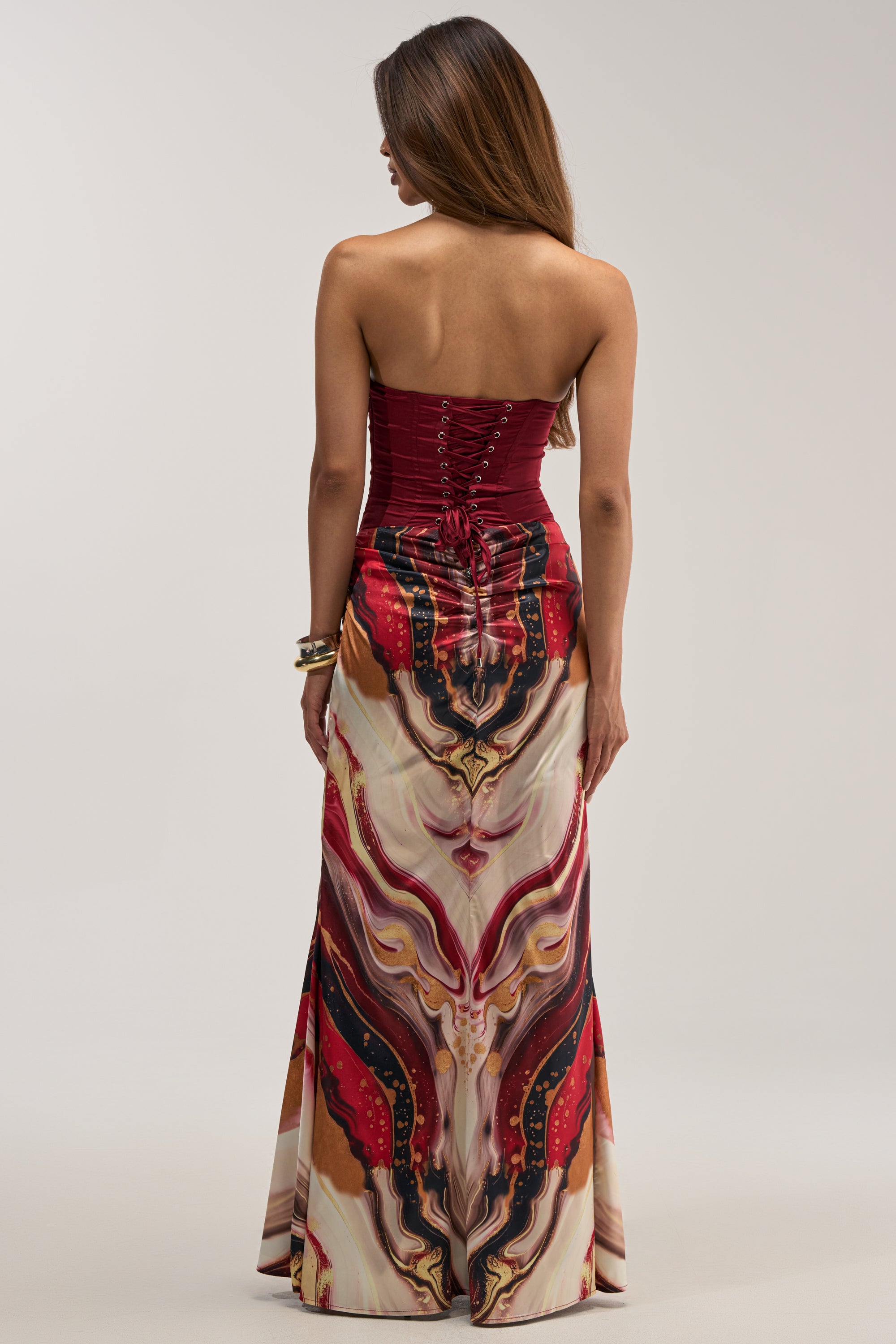 A woman stands with her back to the camera against a plain light background, wearing the SAMARA ABSTRACT MAXI DRESS IN RED MULTI—a strapless burgundy corset maxi dress featuring a long skirt adorned with a vibrant, swirling abstract print.