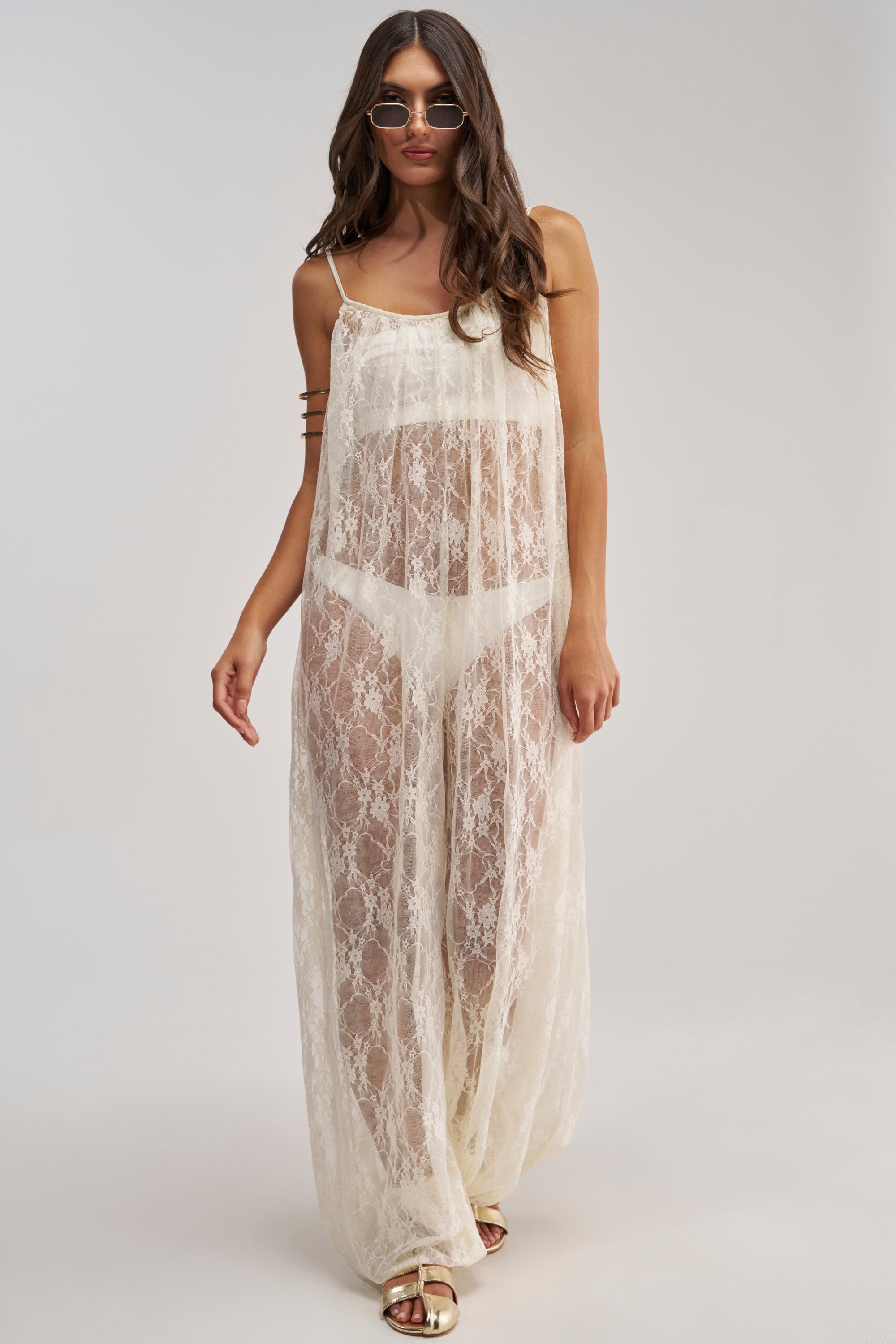 A woman models the MYSTICAL BLOOM LACE JUMPSUIT—a sheer, floor-length white lace piece worn over white undergarments with sunglasses and sandals, highlighting feminine style against a simple, light background.