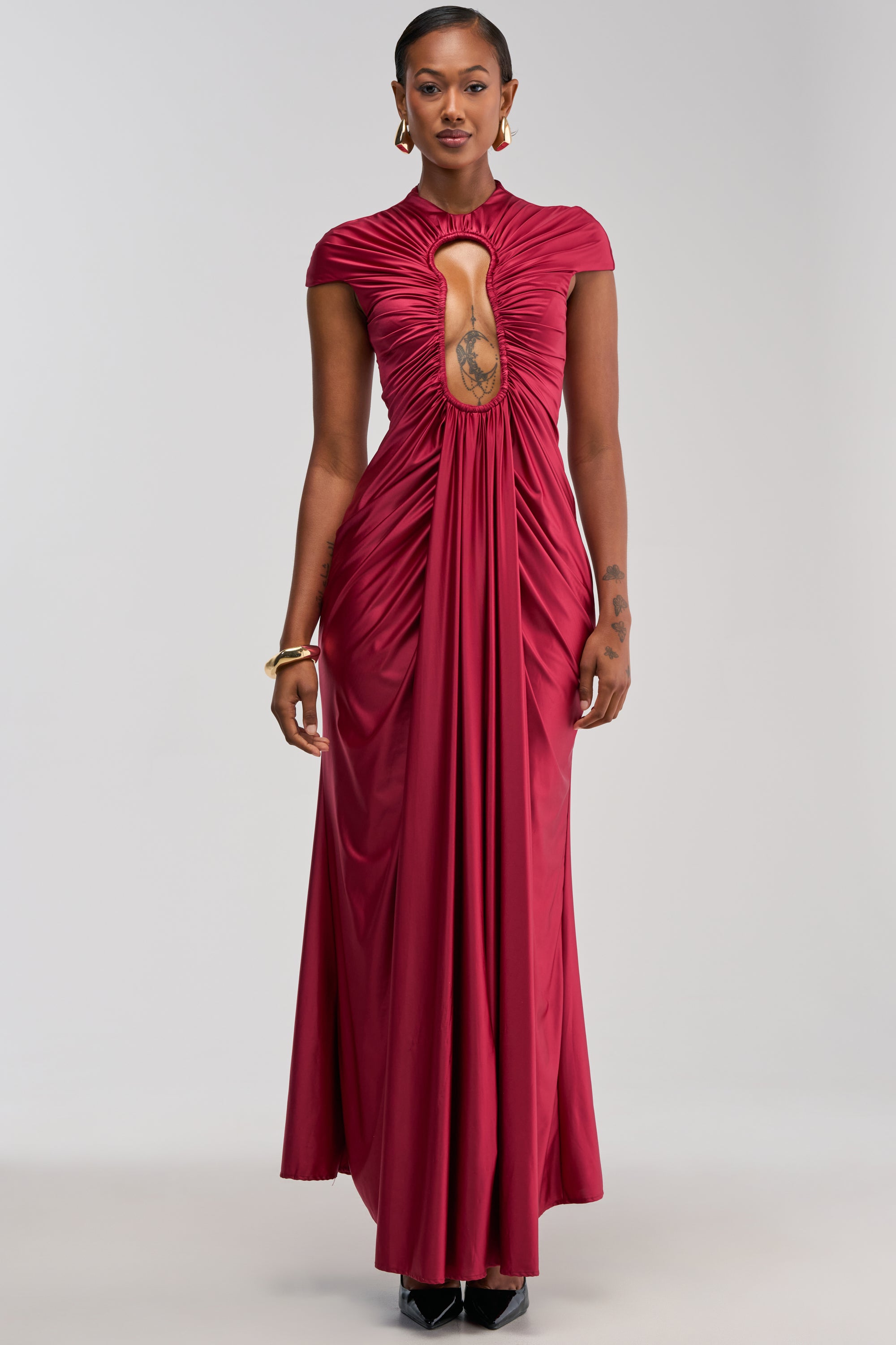 A woman stands facing forward in the Key To My Heart Maxi Dress in Burgundy—a floor-length, short-sleeve gown featuring a gathered keyhole cutout at the chest. She accessorizes with gold jewelry and showcases tattoos on her left arm, making this an elegant choice for a wedding guest look.
