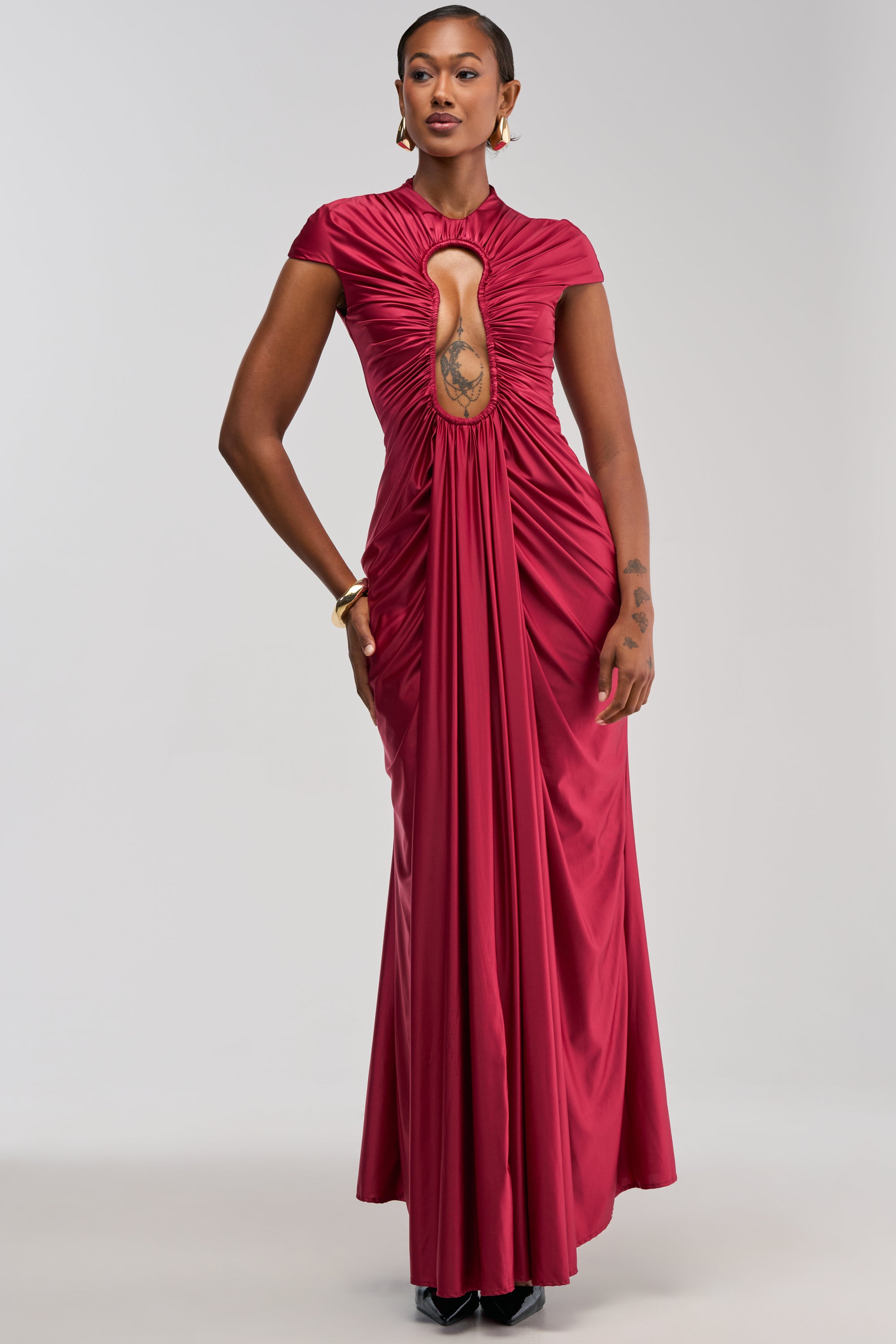 Wearing the KEY TO MY HEART MAXI DRESS IN BURGUNDY, a woman poses in a floor-length silhouette featuring cap sleeves, a gathered lock cutout at the chest, and draped front pleats. She accessorizes with gold earrings and bracelets, standing against a plain light gray background.