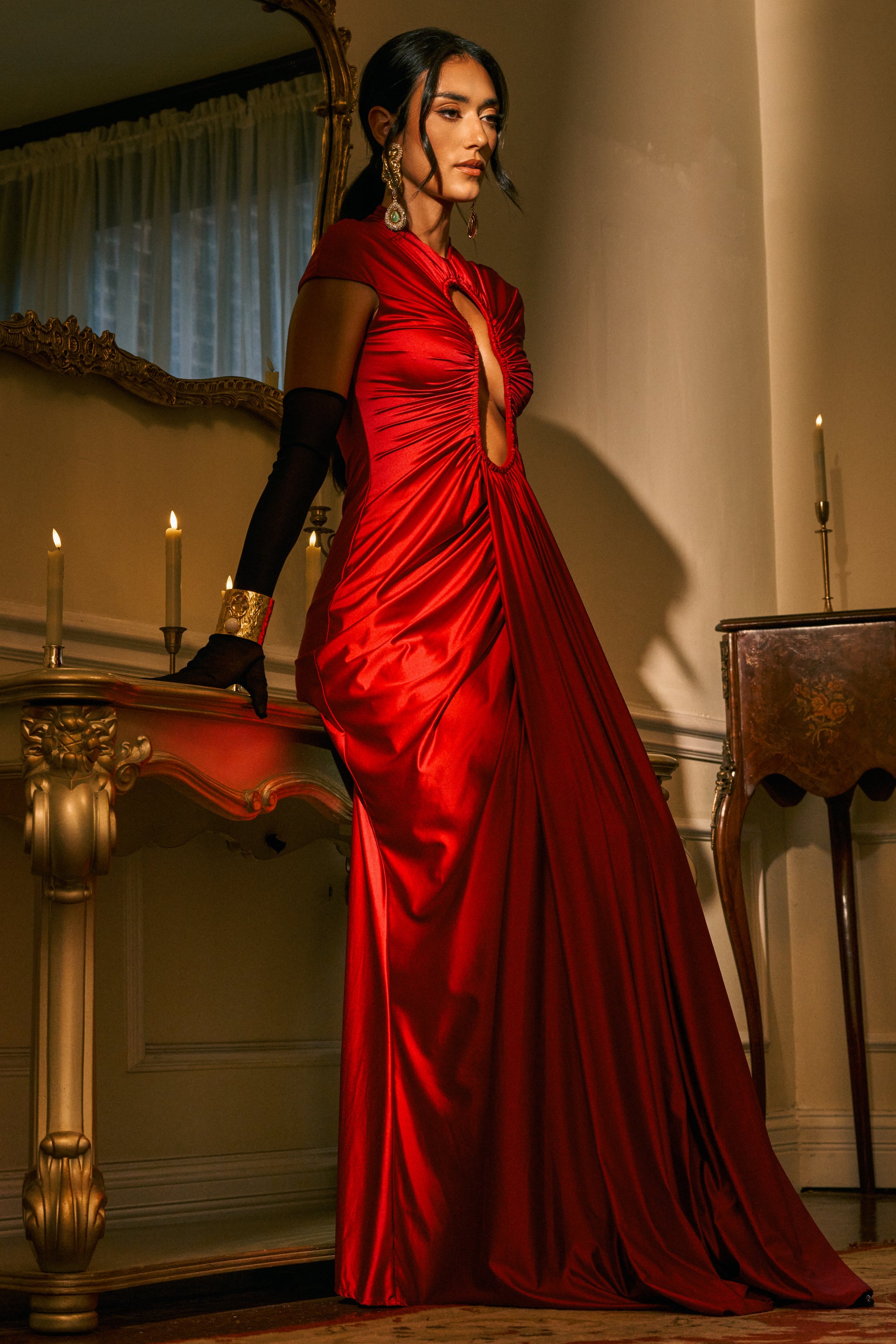 Wearing the KEY TO MY HEART MAXI DRESS IN BURGUNDY—a shimmering maxi dress with a keyhole neckline—a woman stands beside an ornate table adorned with candles. She pairs her look with long black gloves, statement earrings, and a gold bracelet in an elegantly warm and inviting room.