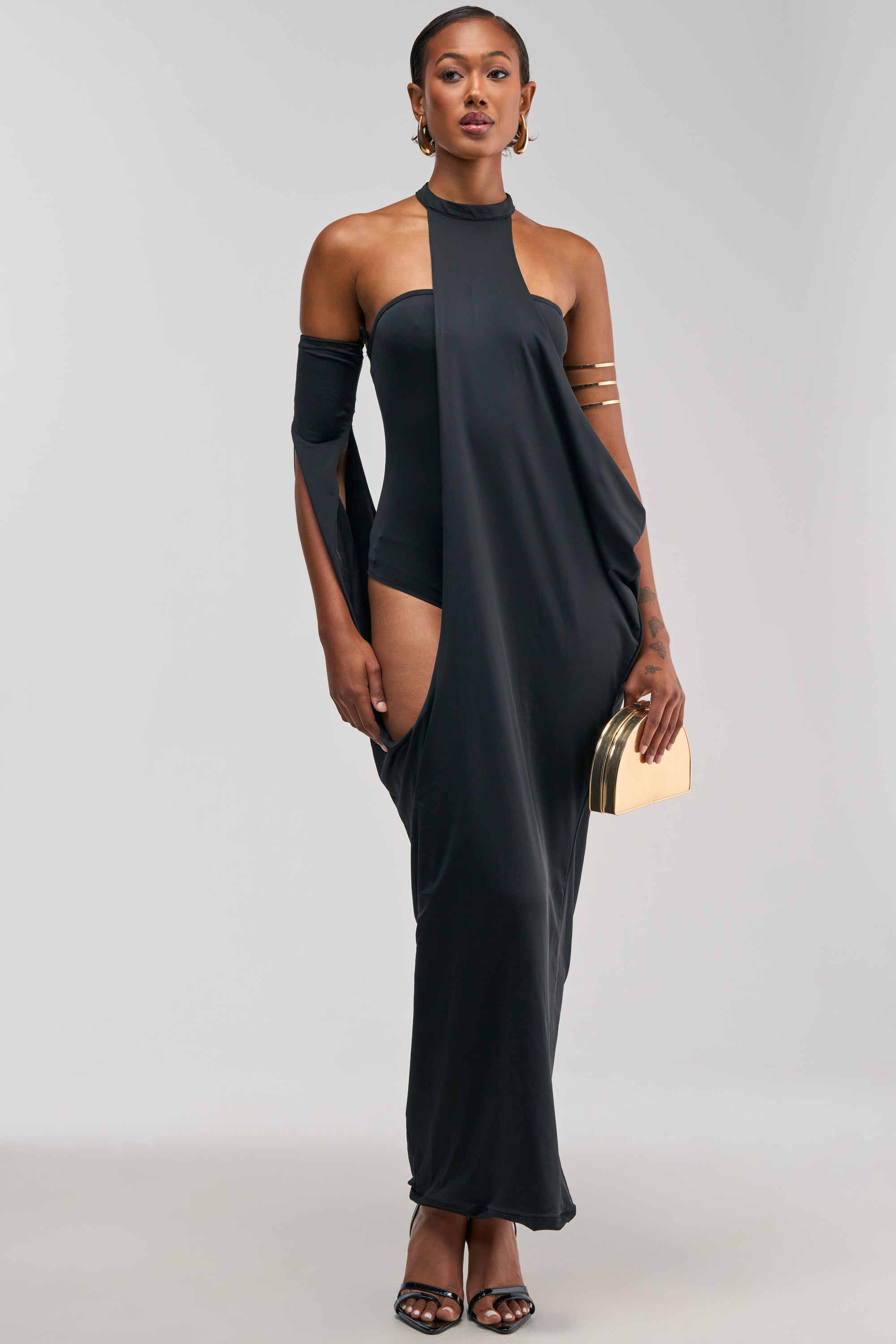 A woman showcases the OPTICAL ILLUSION MAXI DRESS IN BLACK—a unique cutout halter maxi dress with off-the-shoulder sleeves—styled with a gold clutch, heels, earrings, and an arm cuff for a standout special occasion look, set against a plain light background.