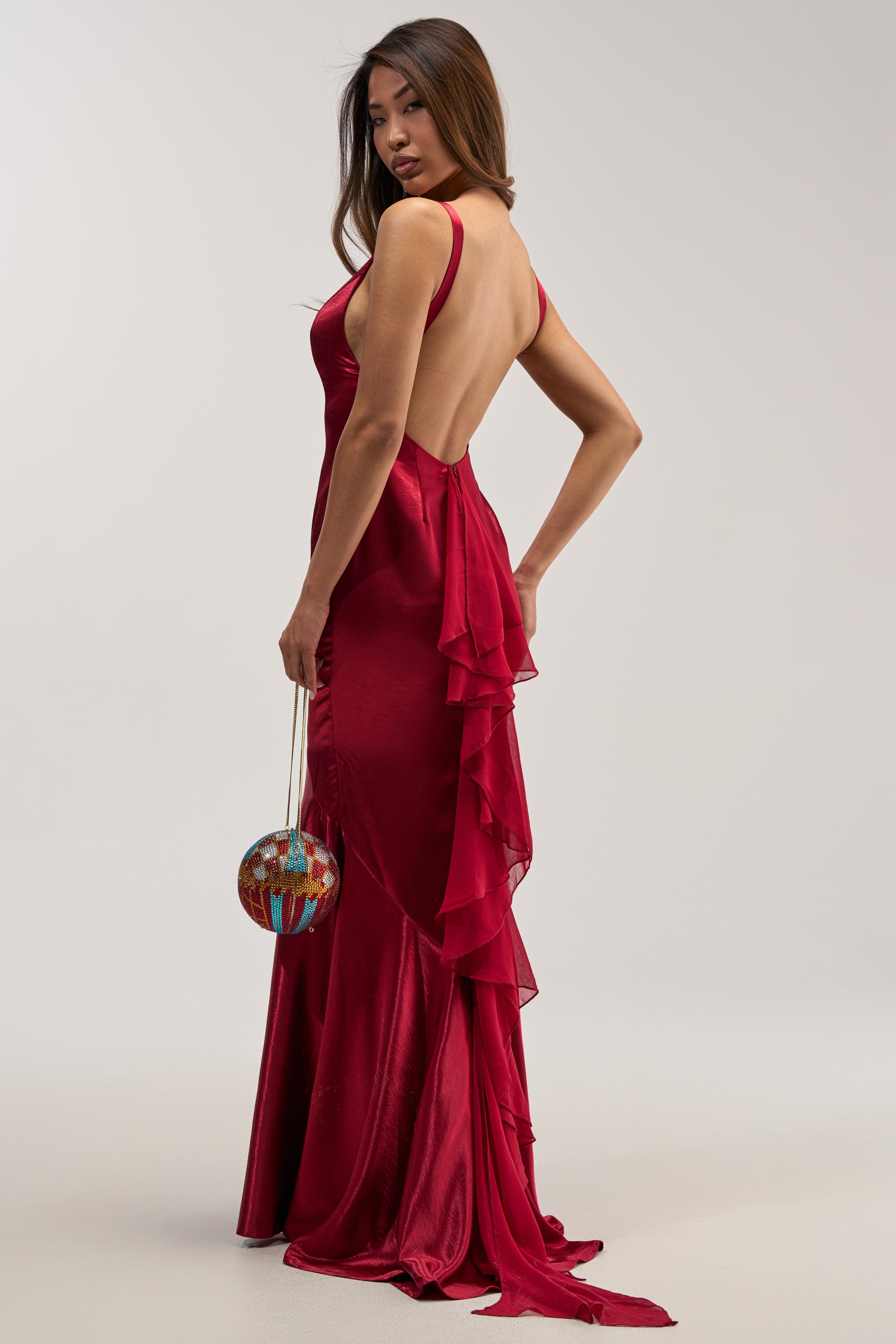 A woman stands on a neutral background, wearing the LUCKY ME LOW BACK MAXI DRESS IN RED—an elegant deep red satin mermaid gown with ruffle details—and holds a round, colorful handbag by her side.