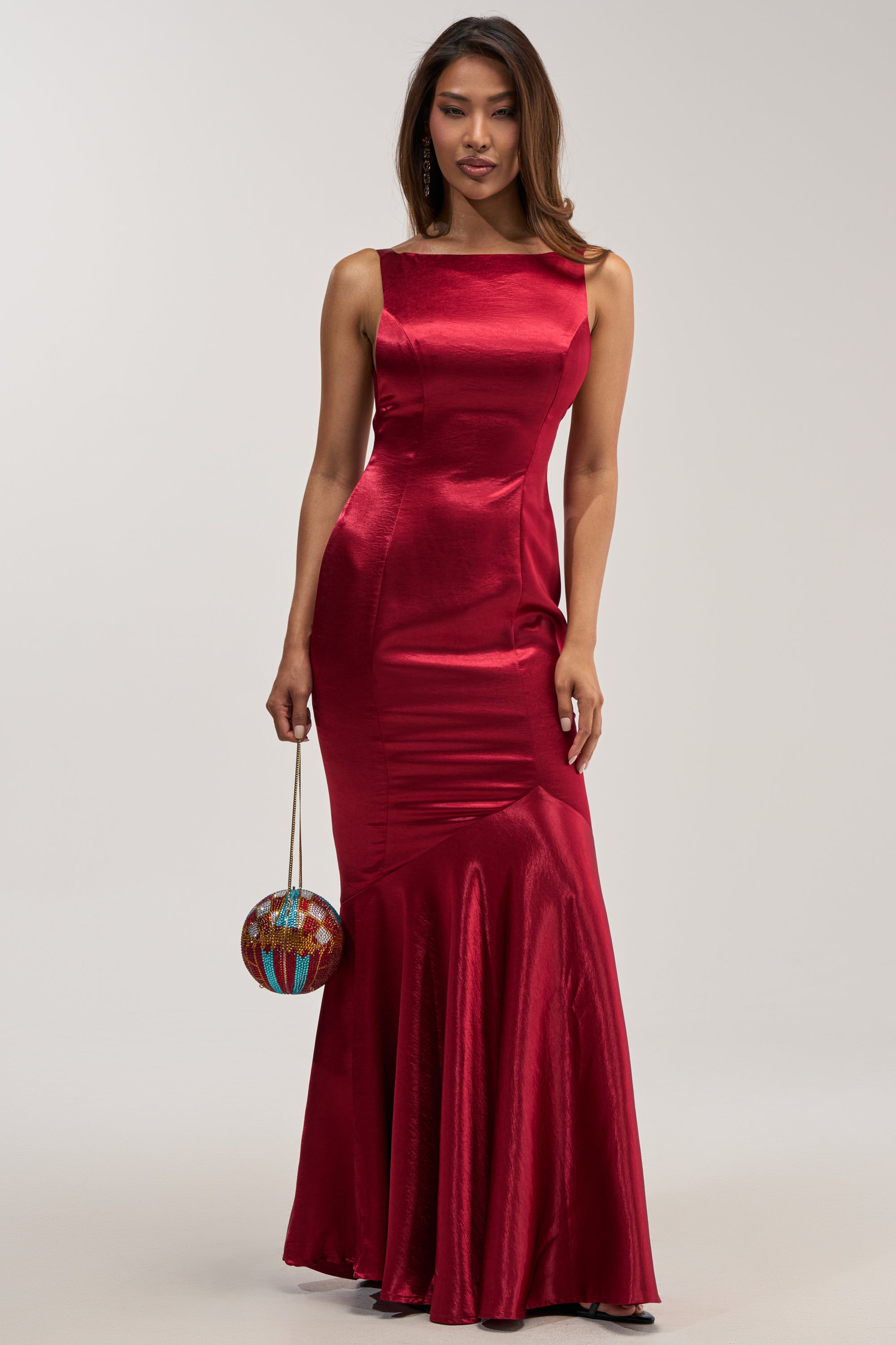 A model with long brown hair poses against a gray backdrop, wearing the LUCKY ME LOW BACK MAXI DRESS IN RED—a sleeveless, floor-length crimson satin mermaid gown. In her right hand, she carries a small, round, multi-colored jeweled sphere handbag with a thin gold chain, completing her elegant formal evening look.