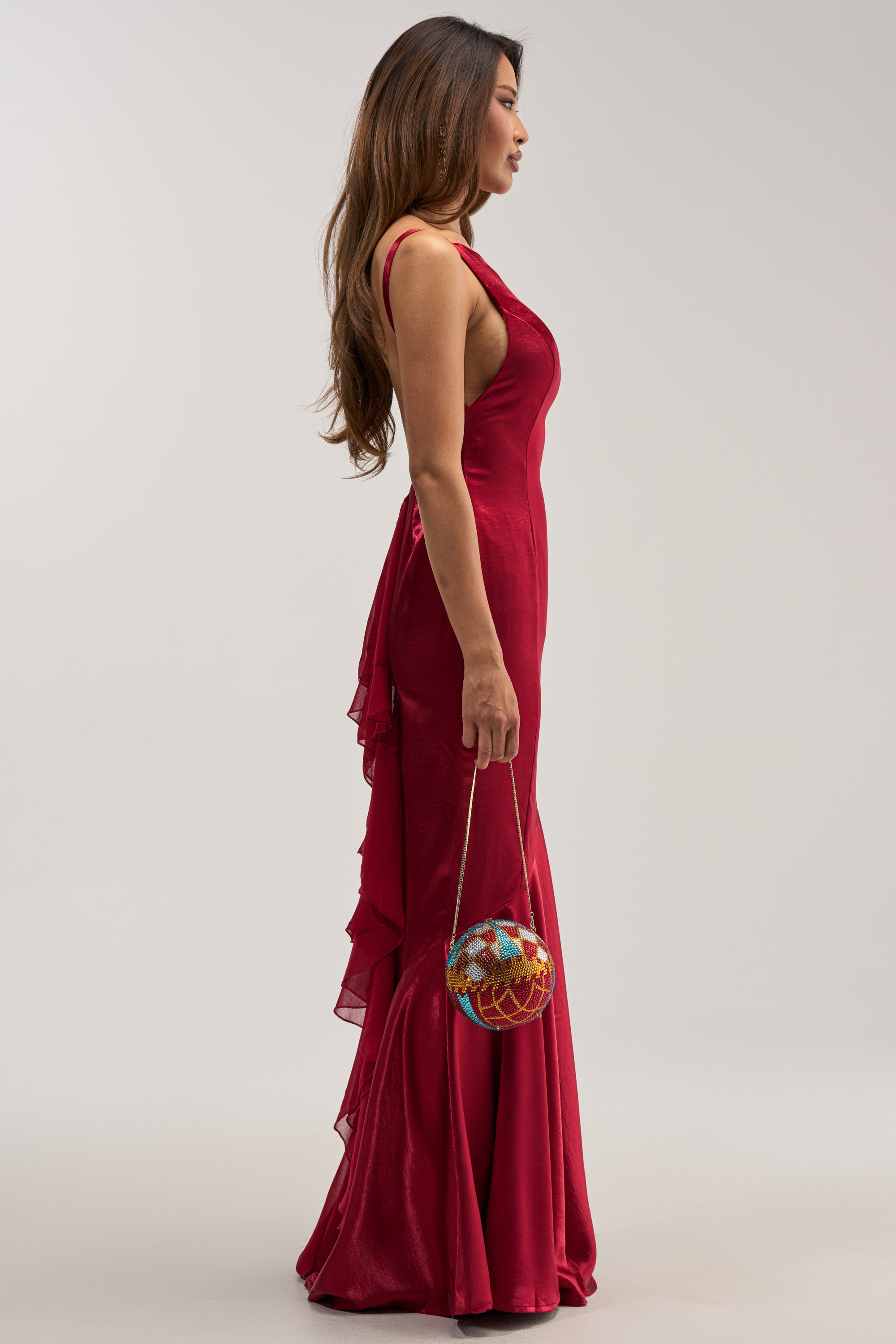 A model with long brown hair, shown in profile against a gray background, wears the floor-length LUCKY ME LOW BACK MAXI DRESS IN RED. The backless satin mermaid gown features ruffles cascading down the back. She carries a small, round handbag adorned with vibrant multicolored rhinestones and suspended from a delicate gold chain.