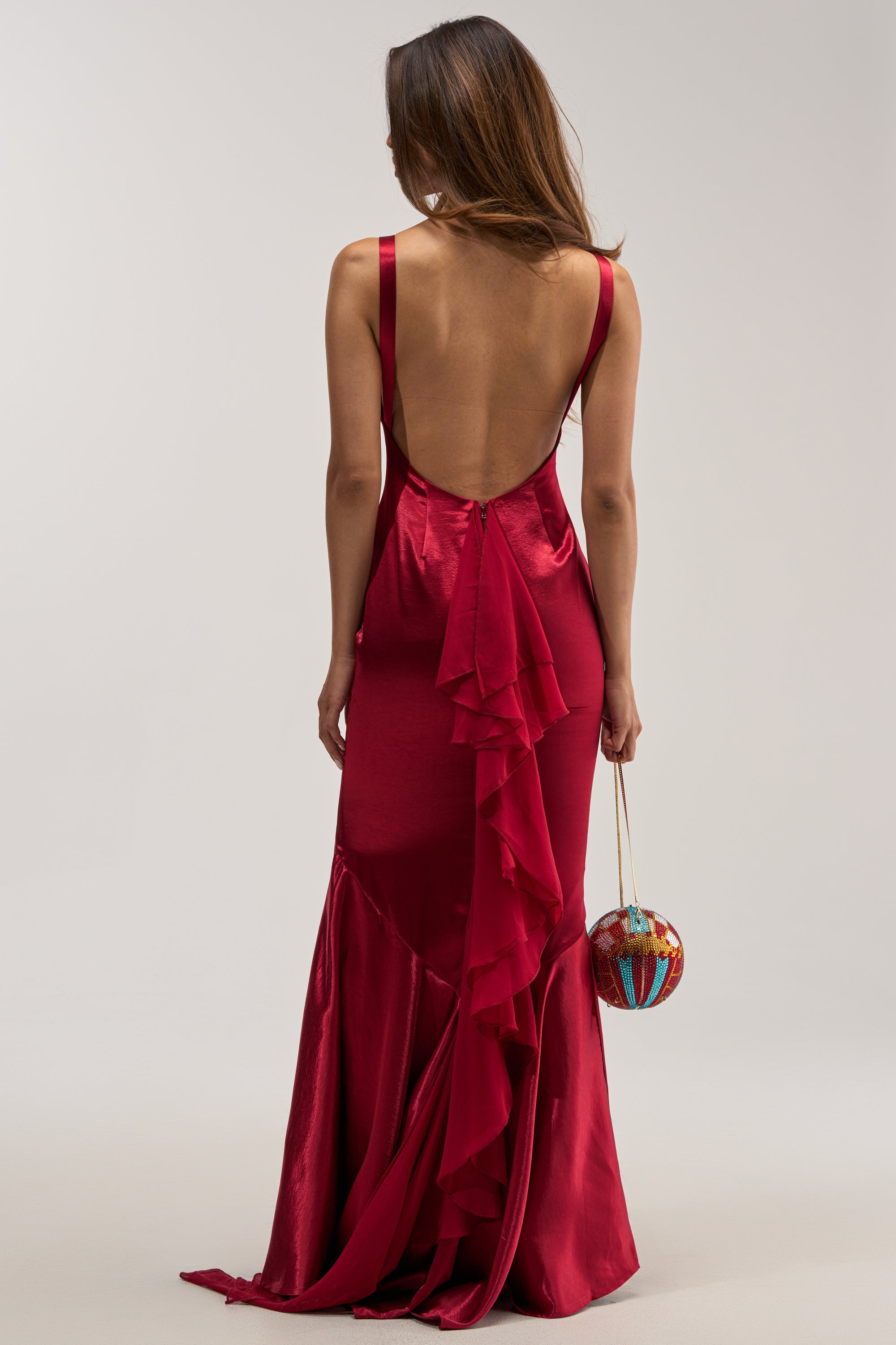 A back view of a woman with long brown hair wearing the LUCKY ME LOW BACK MAXI DRESS IN RED, an elegant, floor-length satin evening gown featuring a stunning low back, delicate spaghetti straps, and a vertical ruffled detail down the back. She is holding a small, round, colorful beaded clutch bag.