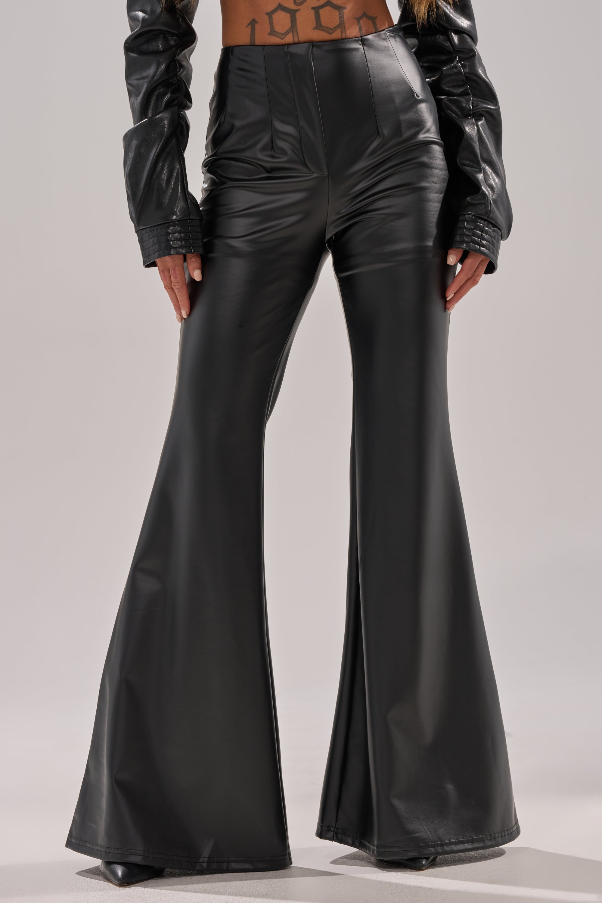 Wearing the DONT CRY BABY GIRL FAUX LEATHER FLARE TROUSER in high-rise black faux leather, paired with a matching black faux leather jacket. Only the lower body is shown against a plain background.