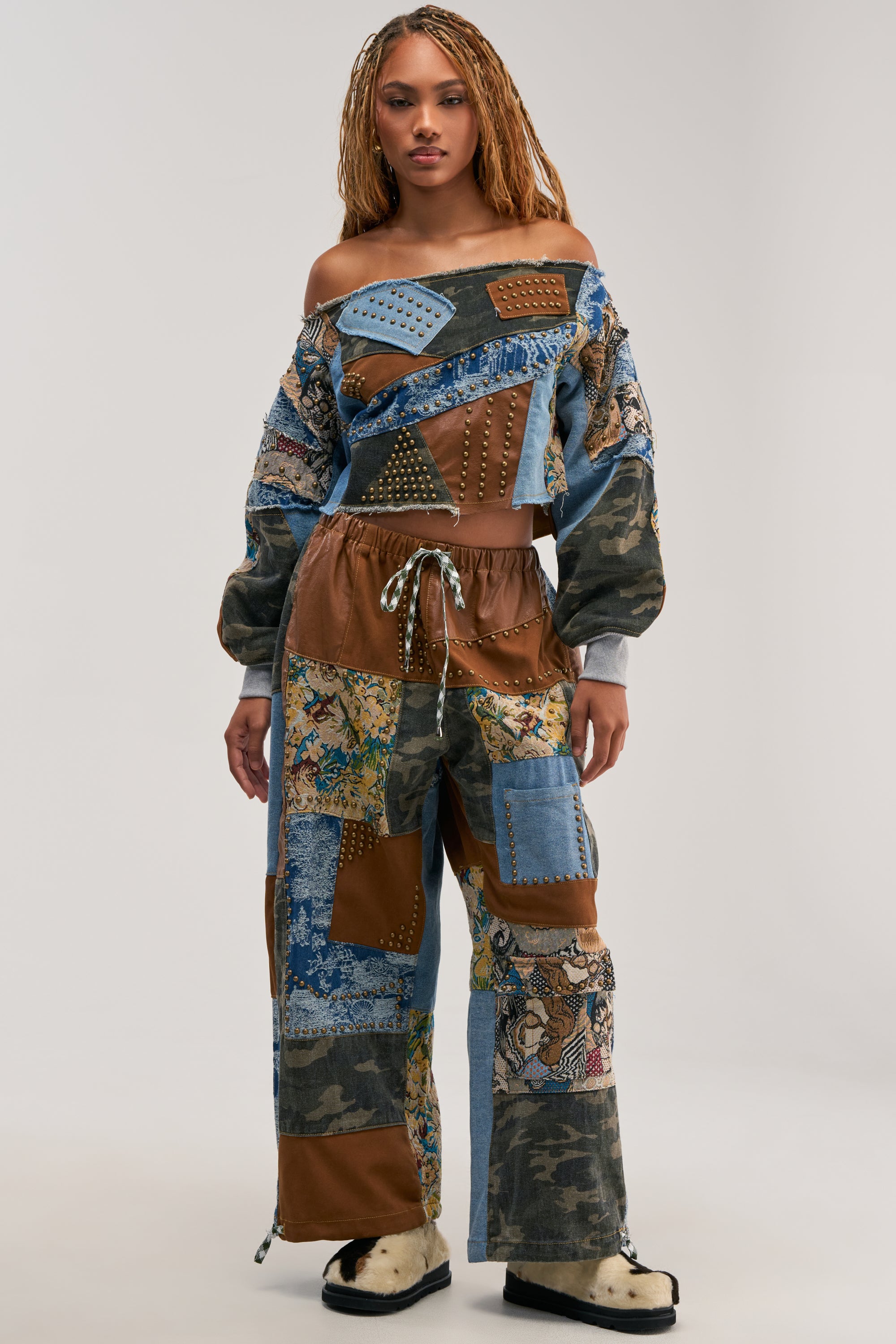 A woman stands facing forward wearing the CALL EM CRAZY PATCHWORK JOGGER IN BROWN MULTI—an eye-catching piece ideal for fall fashion, featuring a mix of denim, camouflage, and leather-patterned fabrics for a trendy streetwear vibe. She completes the look with cream-colored shoes.