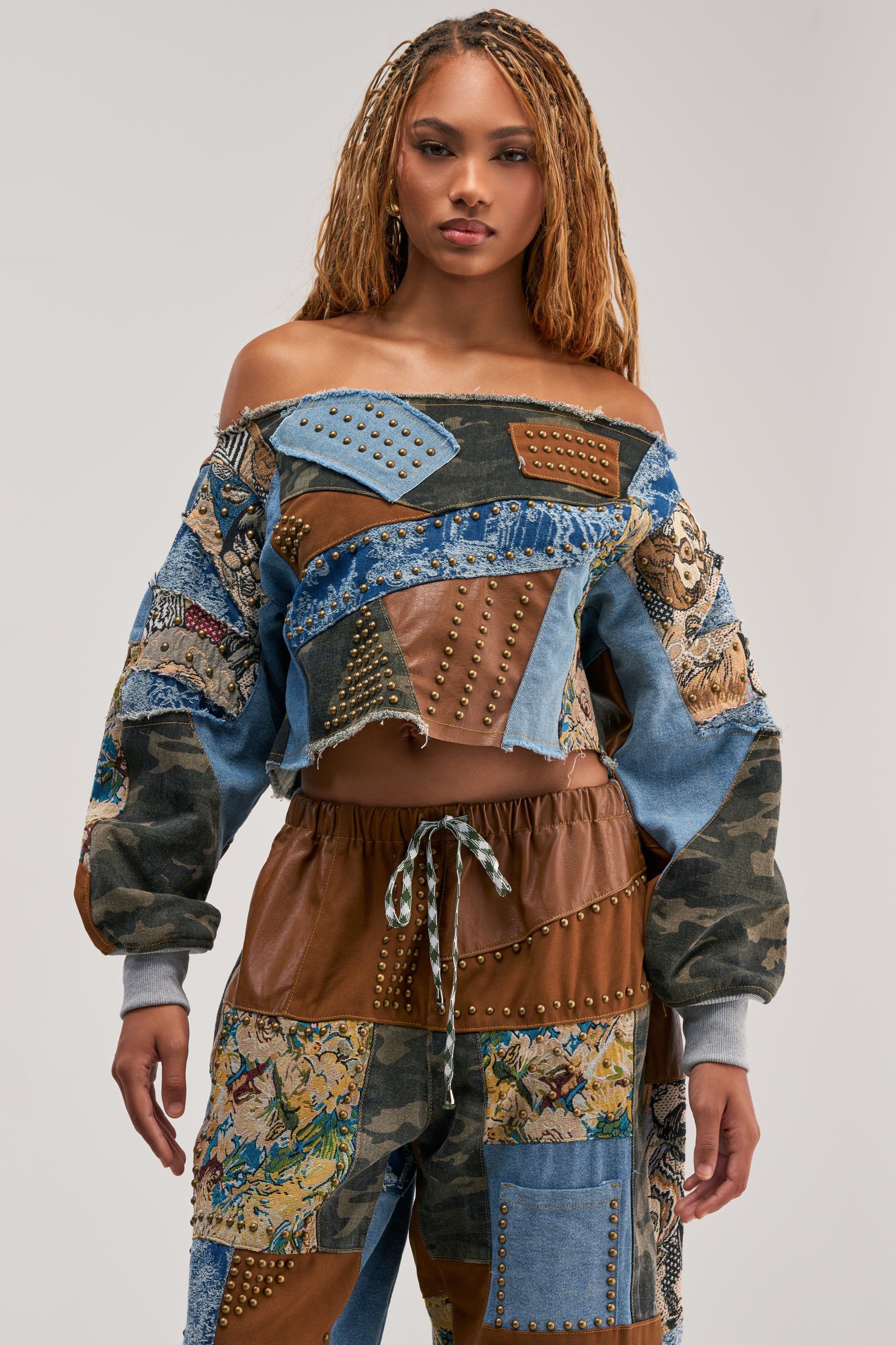 A woman wears the CALL ME CRAZY Patchwork Off the Shoulder Sweatshirt in Brown Multi—an edgy, streetwear-inspired piece featuring denim, brown fabric, and camouflage patches with metal studs and embroidered details. She has long, braided hair and stands against a plain, light background.